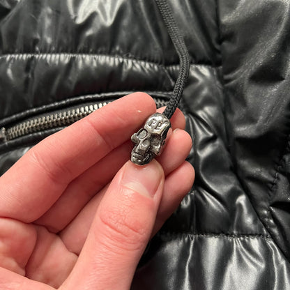 Philipp Plein 2010s Black Waxed Puffer Jacket is a black puffer jacket with a removable fur-trimmed hood attached by zipper. The jacket features heavy silver-tone hardware throughout, including large zipper pulls shaped as crosses with a skull in the center, used on the front pockets and chest pockets. The right chest has a black “P” patch set with crystals, while the left chest features two zip pockets. The hood drawstrings are finished with silver skull tips marked with a “P.”

Both shoulders are finished