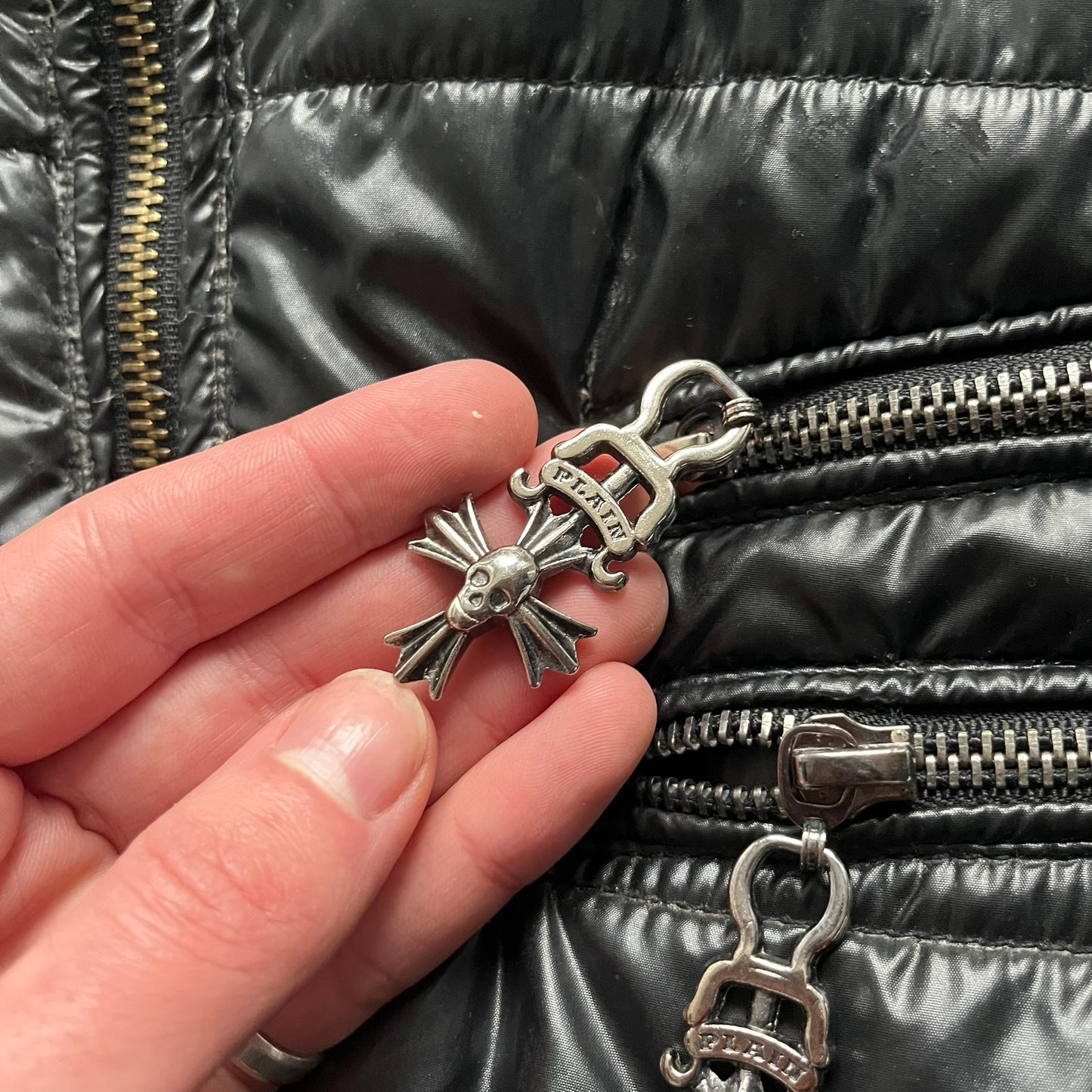 Philipp Plein 2010s Black Waxed Puffer Jacket is a black puffer jacket with a removable fur-trimmed hood attached by zipper. The jacket features heavy silver-tone hardware throughout, including large zipper pulls shaped as crosses with a skull in the center, used on the front pockets and chest pockets. The right chest has a black “P” patch set with crystals, while the left chest features two zip pockets. The hood drawstrings are finished with silver skull tips marked with a “P.”

Both shoulders are finished