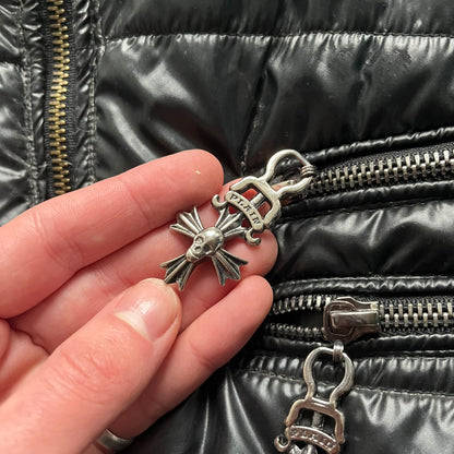 Philipp Plein 2010s Black Waxed Puffer Jacket is a black puffer jacket with a removable fur-trimmed hood attached by zipper. The jacket features heavy silver-tone hardware throughout, including large zipper pulls shaped as crosses with a skull in the center, used on the front pockets and chest pockets. The right chest has a black “P” patch set with crystals, while the left chest features two zip pockets. The hood drawstrings are finished with silver skull tips marked with a “P.”

Both shoulders are finished