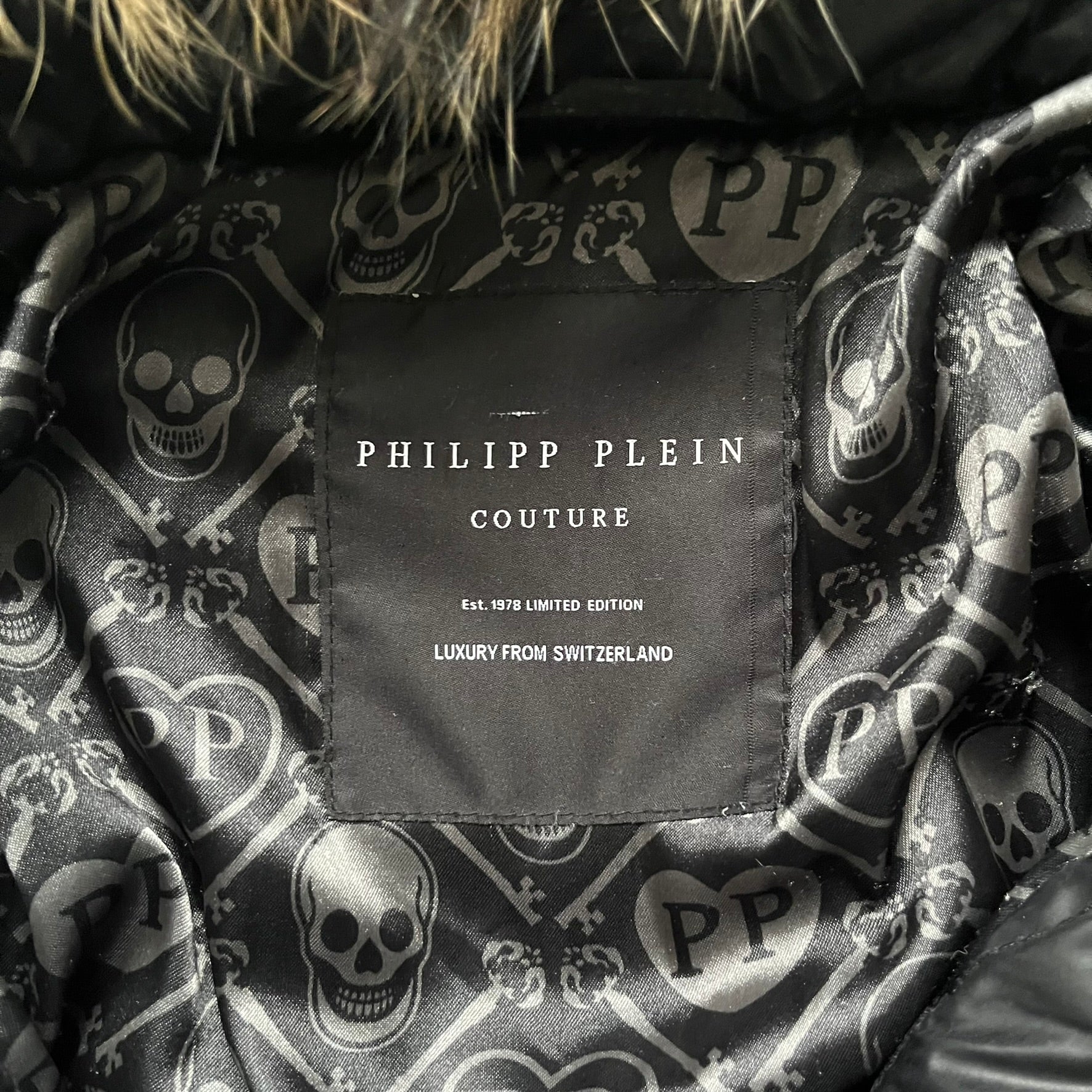 Philipp Plein 2010s Black Waxed Puffer Jacket is a black puffer jacket with a removable fur-trimmed hood attached by zipper. The jacket features heavy silver-tone hardware throughout, including large zipper pulls shaped as crosses with a skull in the center, used on the front pockets and chest pockets. The right chest has a black “P” patch set with crystals, while the left chest features two zip pockets. The hood drawstrings are finished with silver skull tips marked with a “P.”

Both shoulders are finished