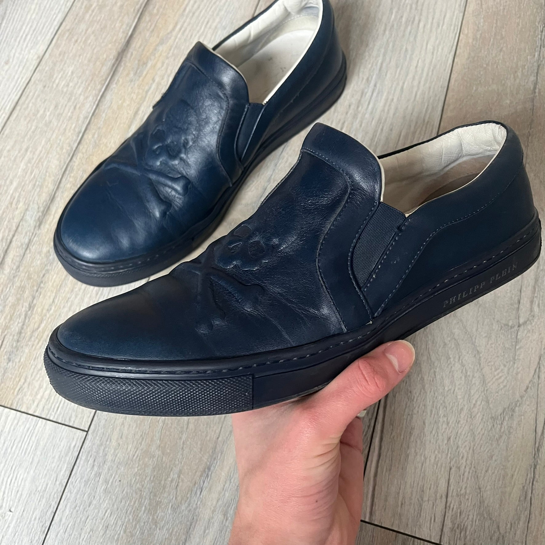 Philipp Plein Navy Skull Slip-Ons are dark navy slip-on sneakers with white interior lining. The front features large raised skull logos, while the sole side displays the Philipp Plein text/logo. Made in Italy, the interior includes an authentic PP tag.