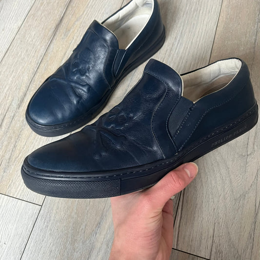 Philipp Plein Navy Skull Slip-Ons are dark navy slip-on sneakers with white interior lining. The front features large raised skull logos, while the sole side displays the Philipp Plein text/logo. Made in Italy, the interior includes an authentic PP tag.