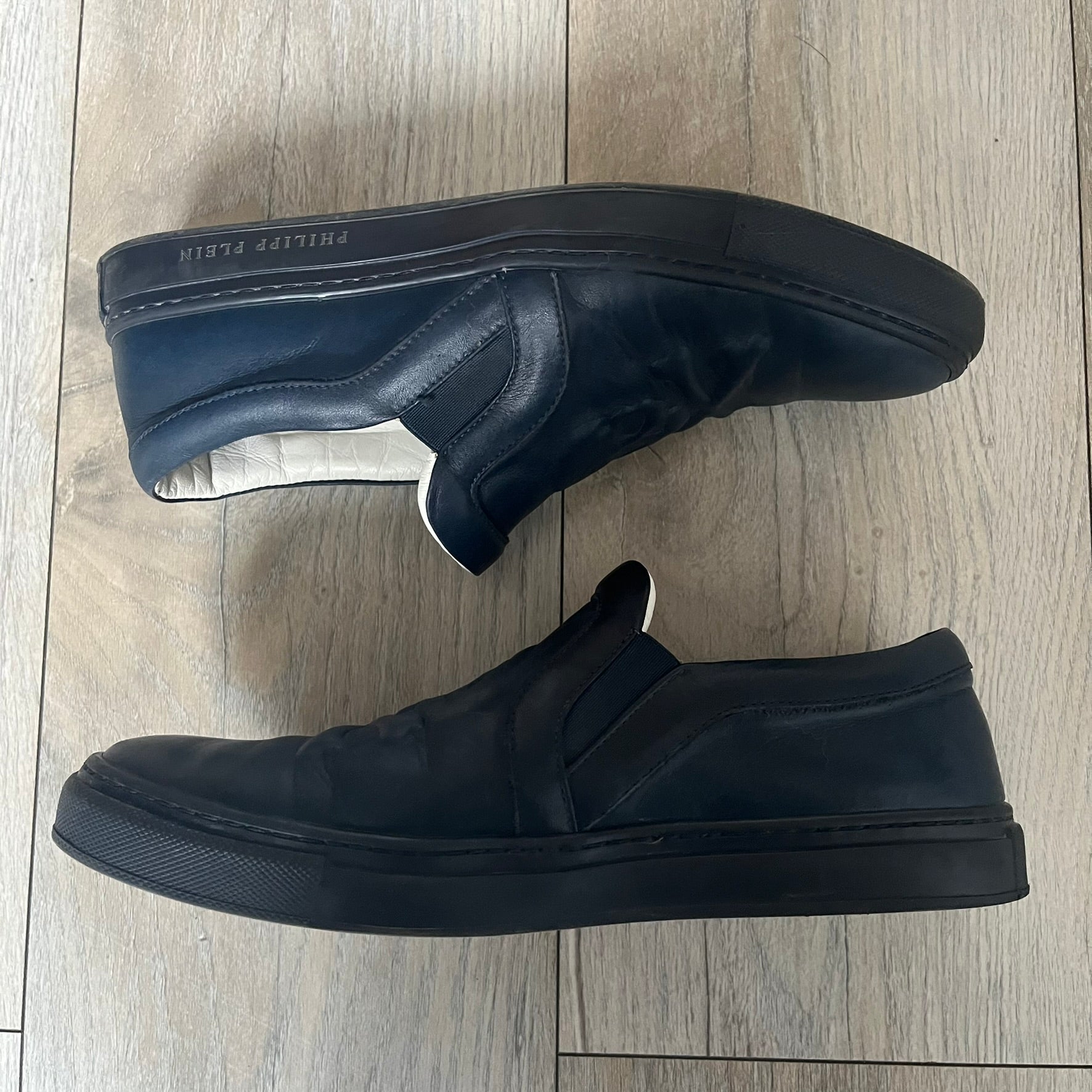 Philipp Plein Navy Skull Slip-Ons are dark navy slip-on sneakers with white interior lining. The front features large raised skull logos, while the sole side displays the Philipp Plein text/logo. Made in Italy, the interior includes an authentic PP tag.