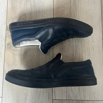 Philipp Plein Navy Skull Slip-Ons are dark navy slip-on sneakers with white interior lining. The front features large raised skull logos, while the sole side displays the Philipp Plein text/logo. Made in Italy, the interior includes an authentic PP tag.