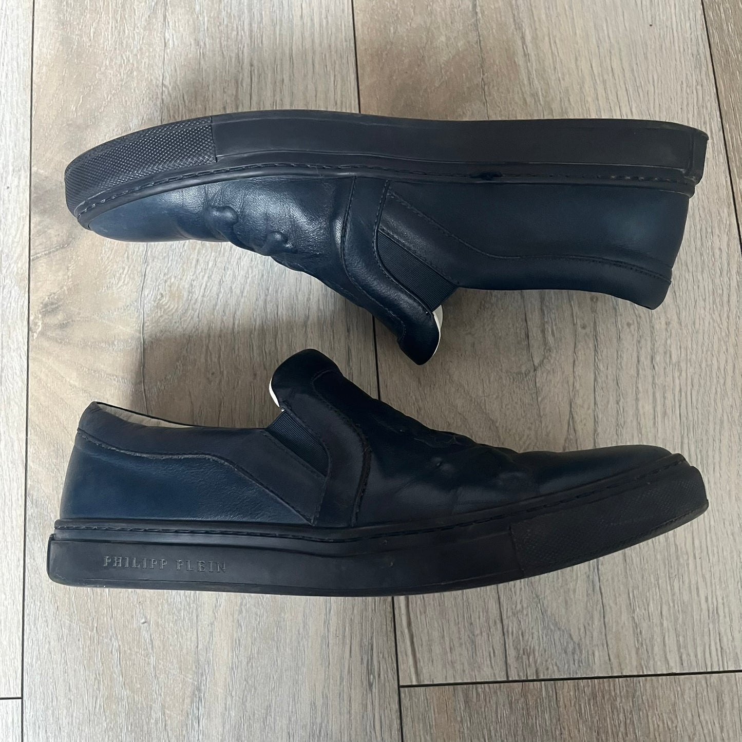 Philipp Plein Navy Skull Slip-Ons are dark navy slip-on sneakers with white interior lining. The front features large raised skull logos, while the sole side displays the Philipp Plein text/logo. Made in Italy, the interior includes an authentic PP tag.