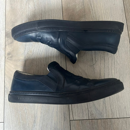 Philipp Plein Navy Skull Slip-Ons are dark navy slip-on sneakers with white interior lining. The front features large raised skull logos, while the sole side displays the Philipp Plein text/logo. Made in Italy, the interior includes an authentic PP tag.