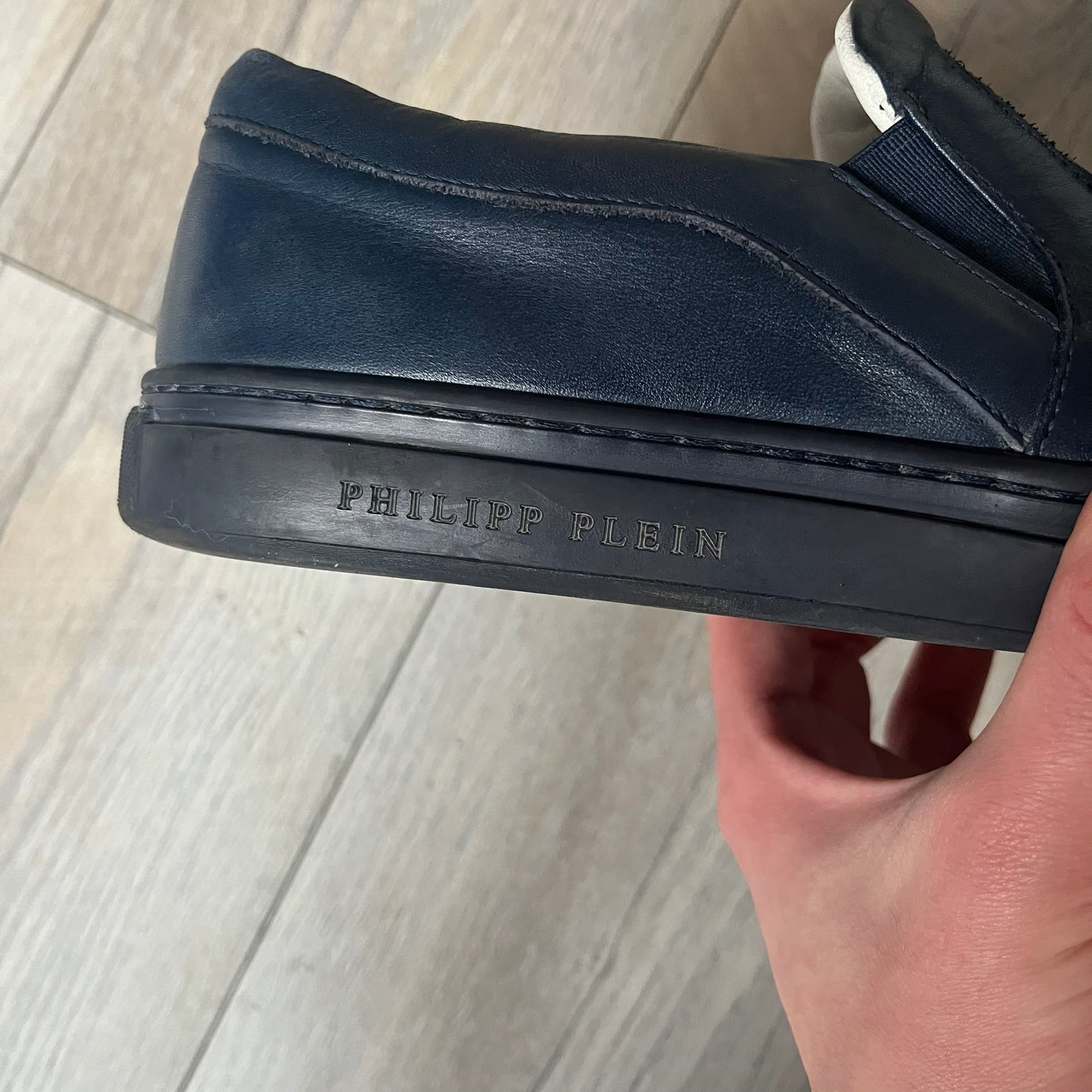 Philipp Plein Navy Skull Slip-Ons are dark navy slip-on sneakers with white interior lining. The front features large raised skull logos, while the sole side displays the Philipp Plein text/logo. Made in Italy, the interior includes an authentic PP tag.