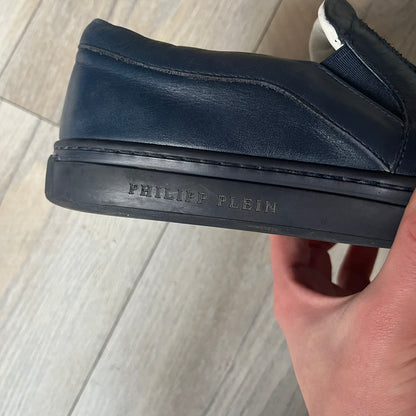 Philipp Plein Navy Skull Slip-Ons are dark navy slip-on sneakers with white interior lining. The front features large raised skull logos, while the sole side displays the Philipp Plein text/logo. Made in Italy, the interior includes an authentic PP tag.