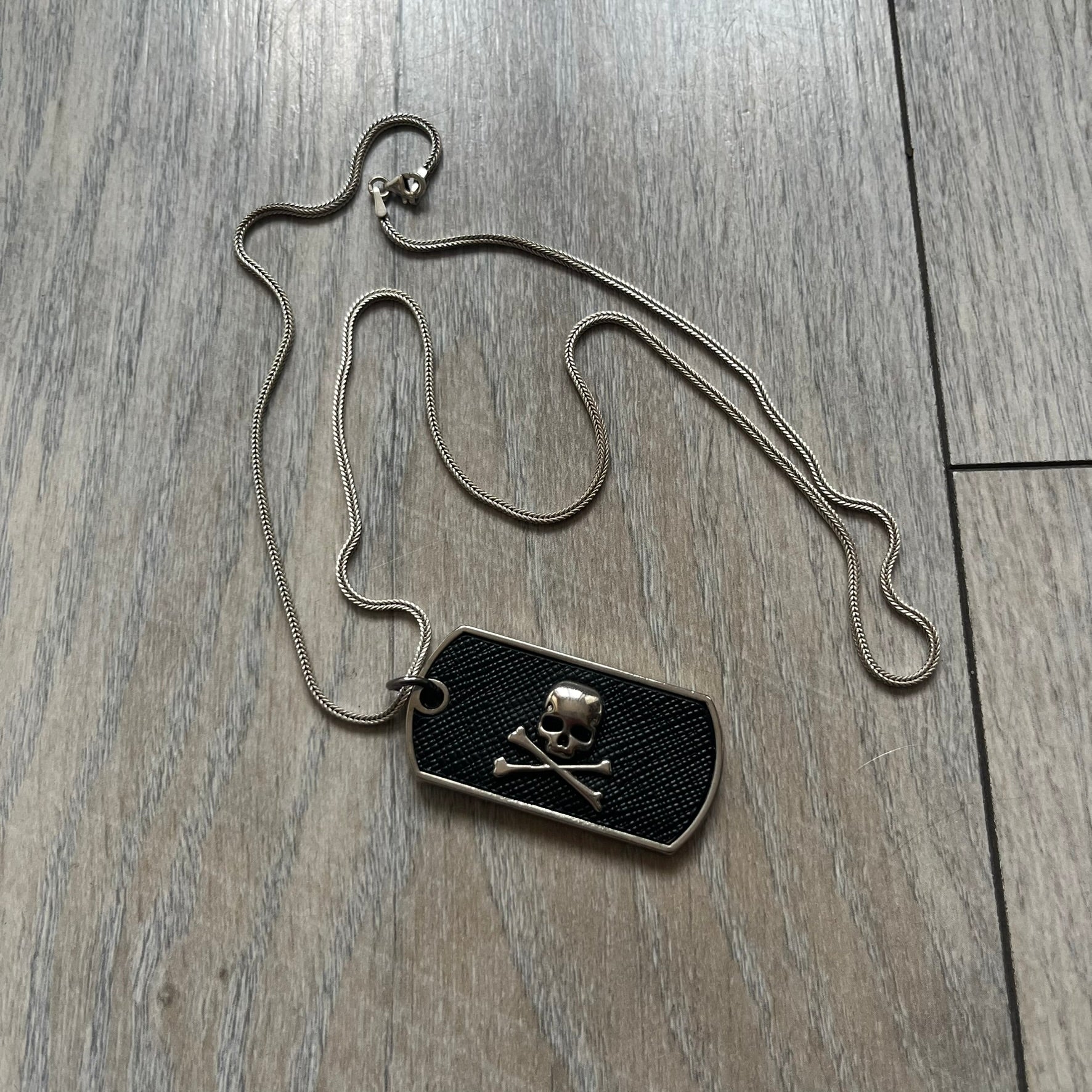 Philipp Plein Skull Dog Tag Necklace features a metal dog tag pendant from Philipp Plein paired with a non-original basic chain. The pendant has a leather inlay on one side with a raised 3D skull and crossbones motif, while the reverse is engraved with the Philipp Plein logo and “Made For” text with a blank line for personalization. Originally used as a zipper pull or accessory attachment for garments or bags, the piece is made in Italy.