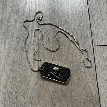 Philipp Plein Skull Dog Tag Necklace features a metal dog tag pendant from Philipp Plein paired with a non-original basic chain. The pendant has a leather inlay on one side with a raised 3D skull and crossbones motif, while the reverse is engraved with the Philipp Plein logo and “Made For” text with a blank line for personalization. Originally used as a zipper pull or accessory attachment for garments or bags, the piece is made in Italy.