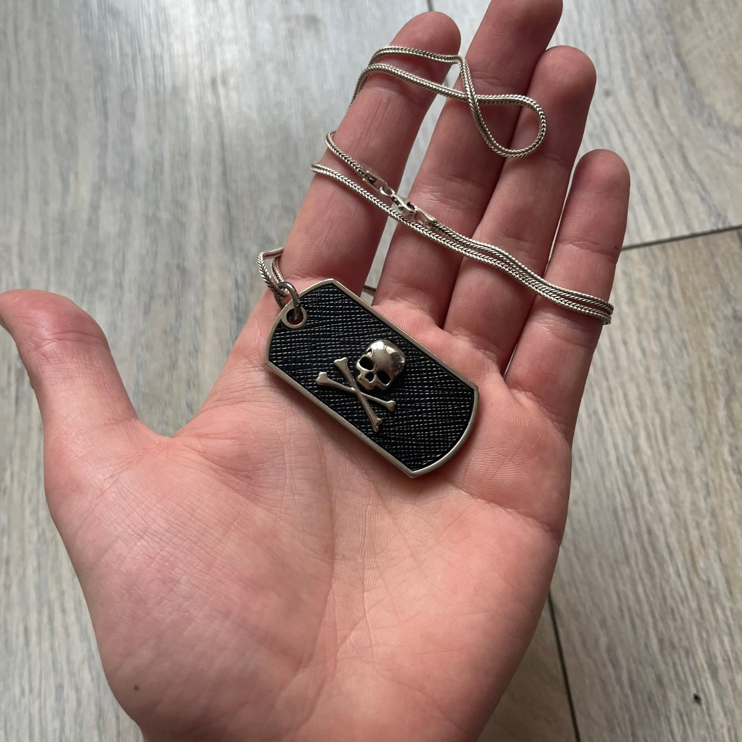 Philipp Plein Skull Dog Tag Necklace features a metal dog tag pendant from Philipp Plein paired with a non-original basic chain. The pendant has a leather inlay on one side with a raised 3D skull and crossbones motif, while the reverse is engraved with the Philipp Plein logo and “Made For” text with a blank line for personalization. Originally used as a zipper pull or accessory attachment for garments or bags, the piece is made in Italy.