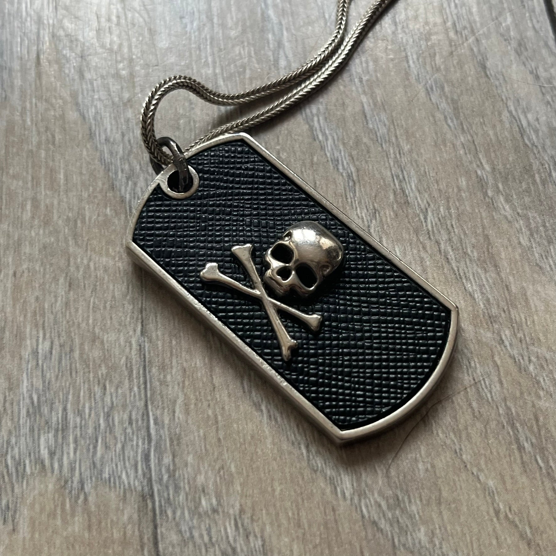 Philipp Plein Skull Dog Tag Necklace features a metal dog tag pendant from Philipp Plein paired with a non-original basic chain. The pendant has a leather inlay on one side with a raised 3D skull and crossbones motif, while the reverse is engraved with the Philipp Plein logo and “Made For” text with a blank line for personalization. Originally used as a zipper pull or accessory attachment for garments or bags, the piece is made in Italy.