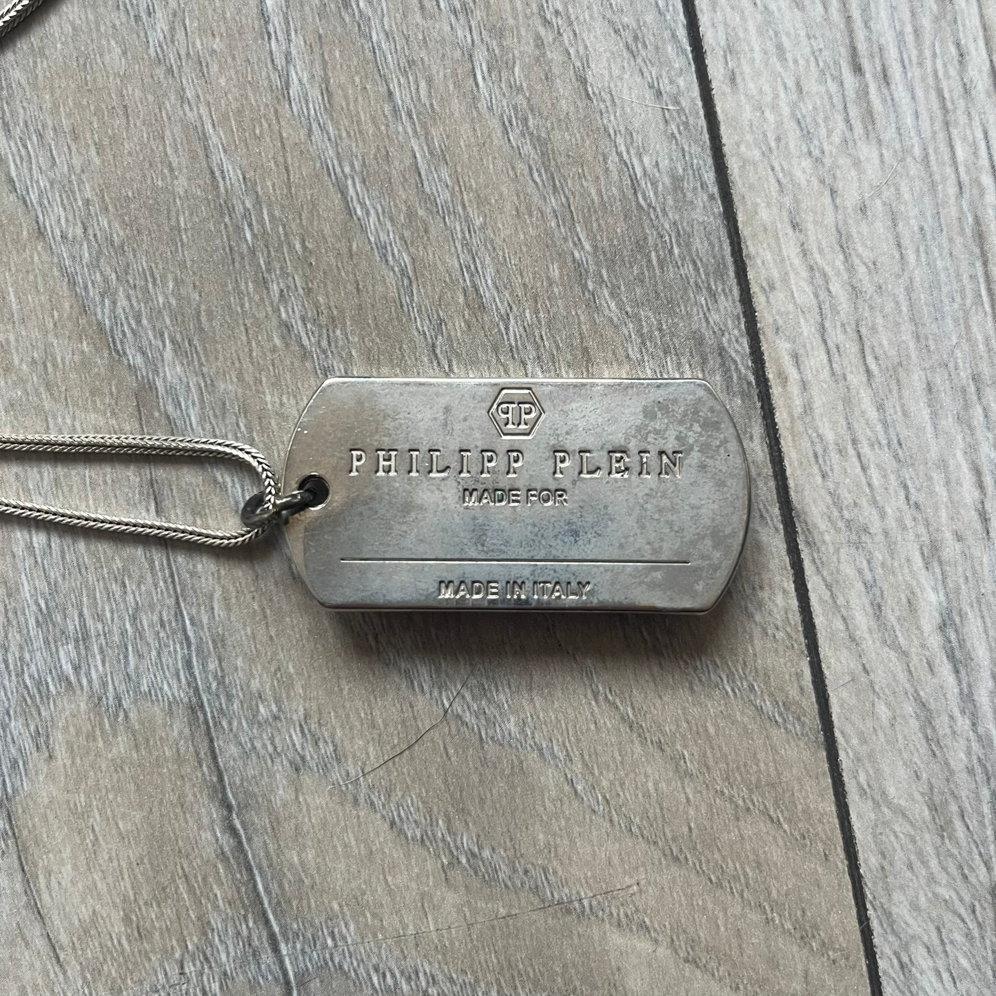 Philipp Plein Skull Dog Tag Necklace features a metal dog tag pendant from Philipp Plein paired with a non-original basic chain. The pendant has a leather inlay on one side with a raised 3D skull and crossbones motif, while the reverse is engraved with the Philipp Plein logo and “Made For” text with a blank line for personalization. Originally used as a zipper pull or accessory attachment for garments or bags, the piece is made in Italy.