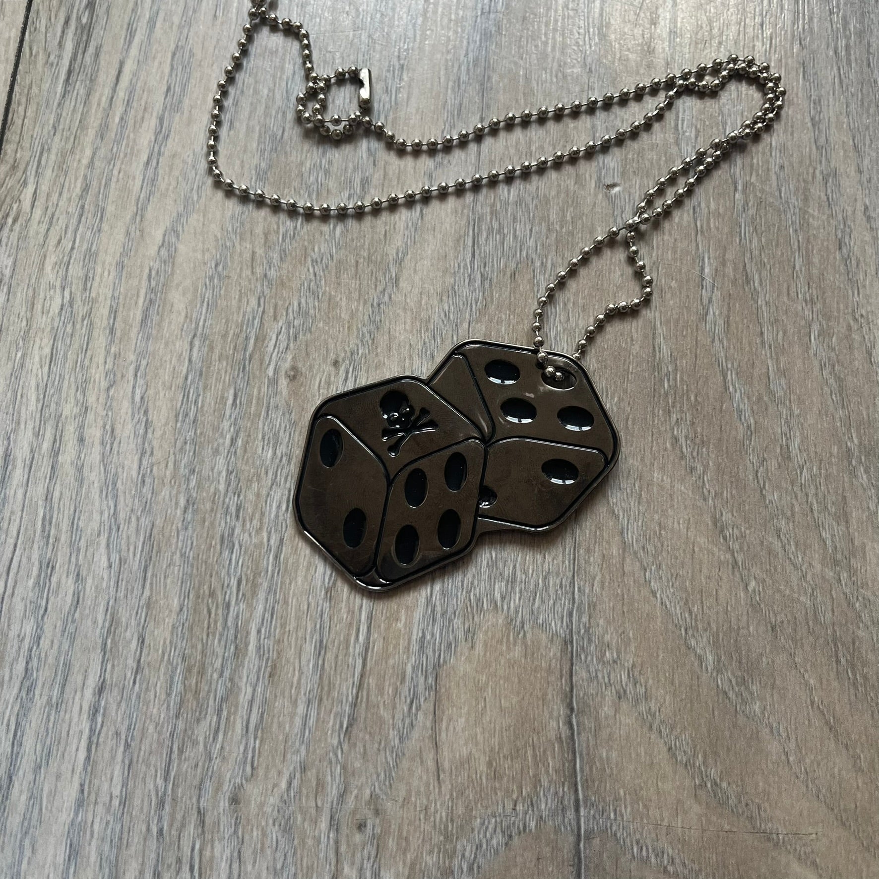 Philipp Plein Two Dices Necklace features a flat metal pendant from Philipp Plein paired with a non-original ball chain. The pendant is designed as two connected dice in a dark grey finish with black enamel dots, with one side replacing the dots with the Philipp Plein skull motif, while the reverse is engraved with the brand logo. Originally used as a zipper pull or accessory attachment, the piece is made in Italy and comes from the 2013 “Hardcore Diamonds” collection.