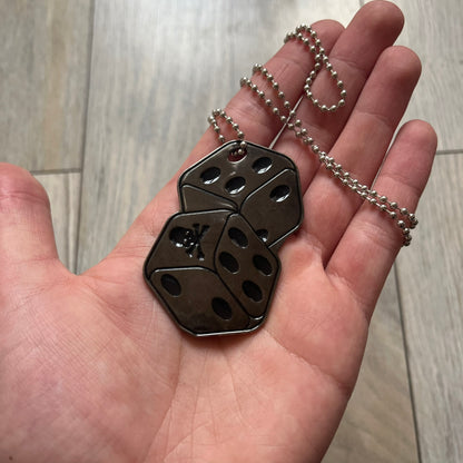 Philipp Plein Two Dices Necklace features a flat metal pendant from Philipp Plein paired with a non-original ball chain. The pendant is designed as two connected dice in a dark grey finish with black enamel dots, with one side replacing the dots with the Philipp Plein skull motif, while the reverse is engraved with the brand logo. Originally used as a zipper pull or accessory attachment, the piece is made in Italy and comes from the 2013 “Hardcore Diamonds” collection.