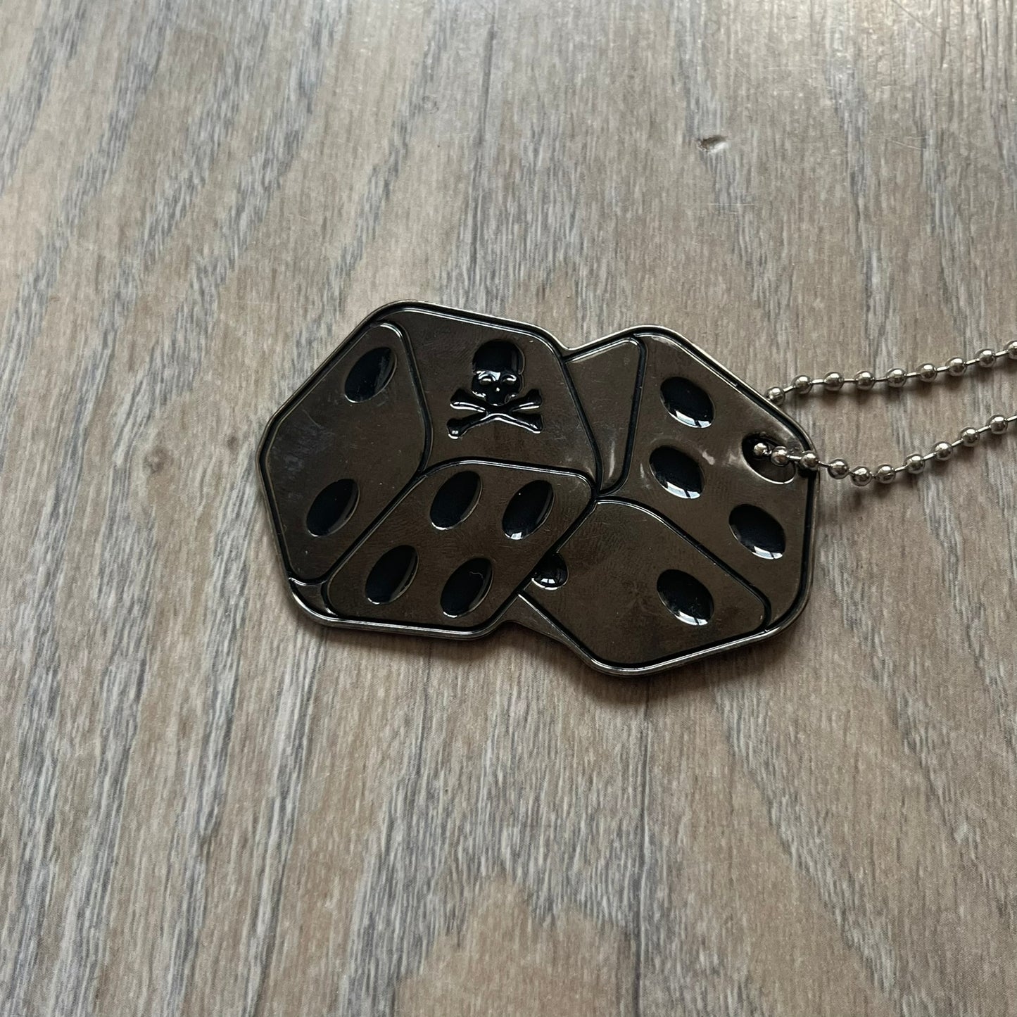 Philipp Plein Two Dices Necklace features a flat metal pendant from Philipp Plein paired with a non-original ball chain. The pendant is designed as two connected dice in a dark grey finish with black enamel dots, with one side replacing the dots with the Philipp Plein skull motif, while the reverse is engraved with the brand logo. Originally used as a zipper pull or accessory attachment, the piece is made in Italy and comes from the 2013 “Hardcore Diamonds” collection.