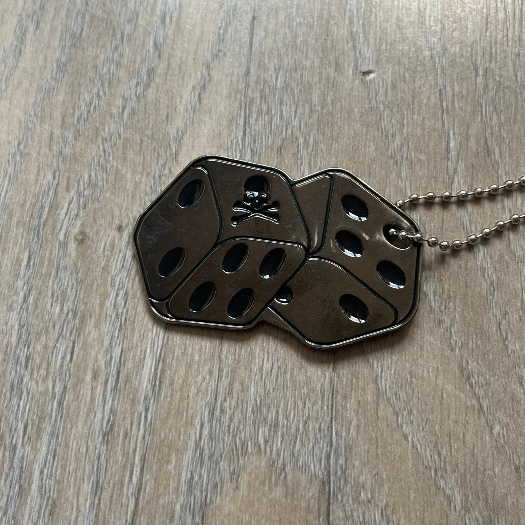 Philipp Plein Two Dices Necklace features a flat metal pendant from Philipp Plein paired with a non-original ball chain. The pendant is designed as two connected dice in a dark grey finish with black enamel dots, with one side replacing the dots with the Philipp Plein skull motif, while the reverse is engraved with the brand logo. Originally used as a zipper pull or accessory attachment, the piece is made in Italy and comes from the 2013 “Hardcore Diamonds” collection.