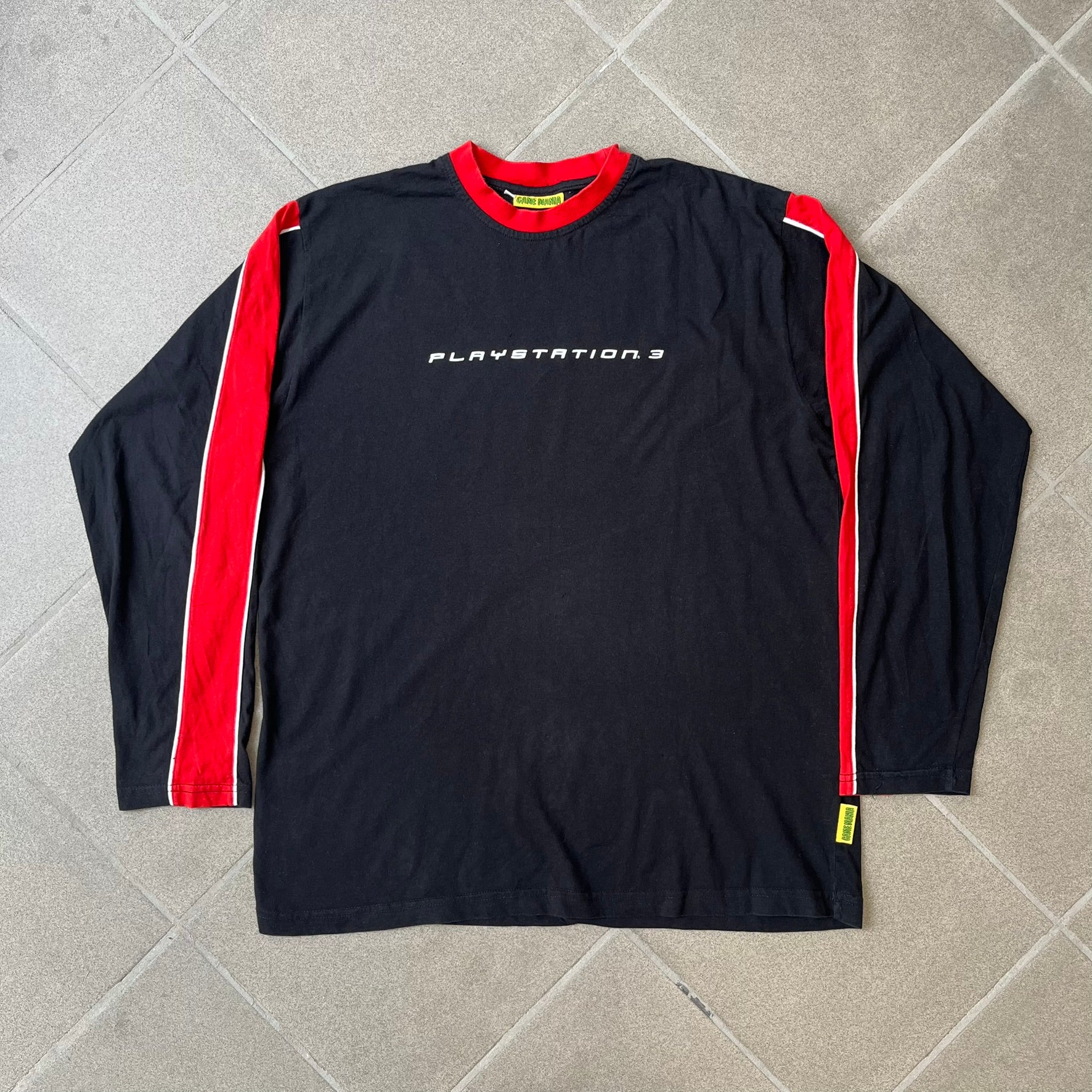 PlayStation 3 Game Mania Crew 2000s Longsleeve is a black long-sleeve shirt featuring red collar and shoulder stripe detailing running along the tops of the sleeves. The front displays a white PlayStation 3 logo, while the back is printed with the "Game Mania" logo above "CREW" lettering. Finished with distinctive yellow Game Mania neck tag, this event/store-exclusive piece was produced for Game Mania staff and promotional use.