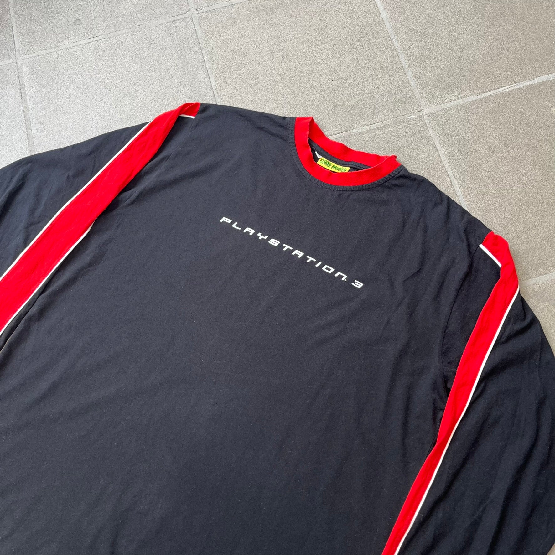 PlayStation 3 Game Mania Crew 2000s Longsleeve is a black long-sleeve shirt featuring red collar and shoulder stripe detailing running along the tops of the sleeves. The front displays a white PlayStation 3 logo, while the back is printed with the "Game Mania" logo above "CREW" lettering. Finished with distinctive yellow Game Mania neck tag, this event/store-exclusive piece was produced for Game Mania staff and promotional use.