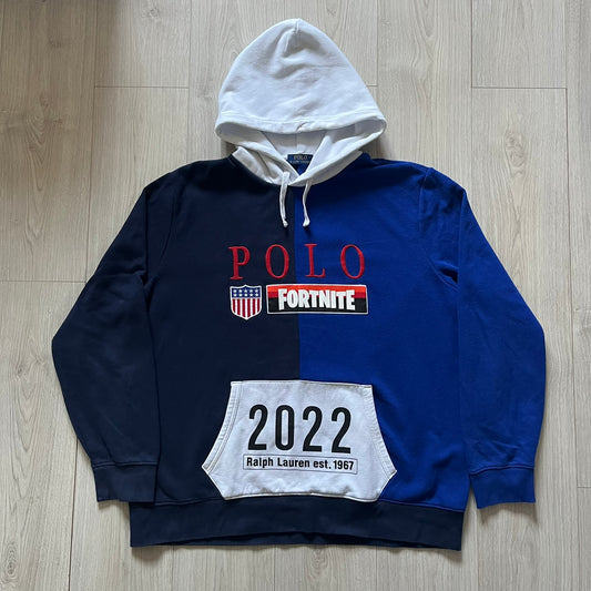 Polo Ralph Lauren x Fortnite 2022 Hoodie featuring a two-tone split design in blue and navy with a white hood. The front showcases a large white panel with “2022” text, Ralph Lauren Est. 1967 branding, and a Fortnite logo, along with an oversized embroidered Polo logo above. A limited collaboration piece combining Ralph Lauren’s heritage aesthetic with Fortnite’s playful edge.