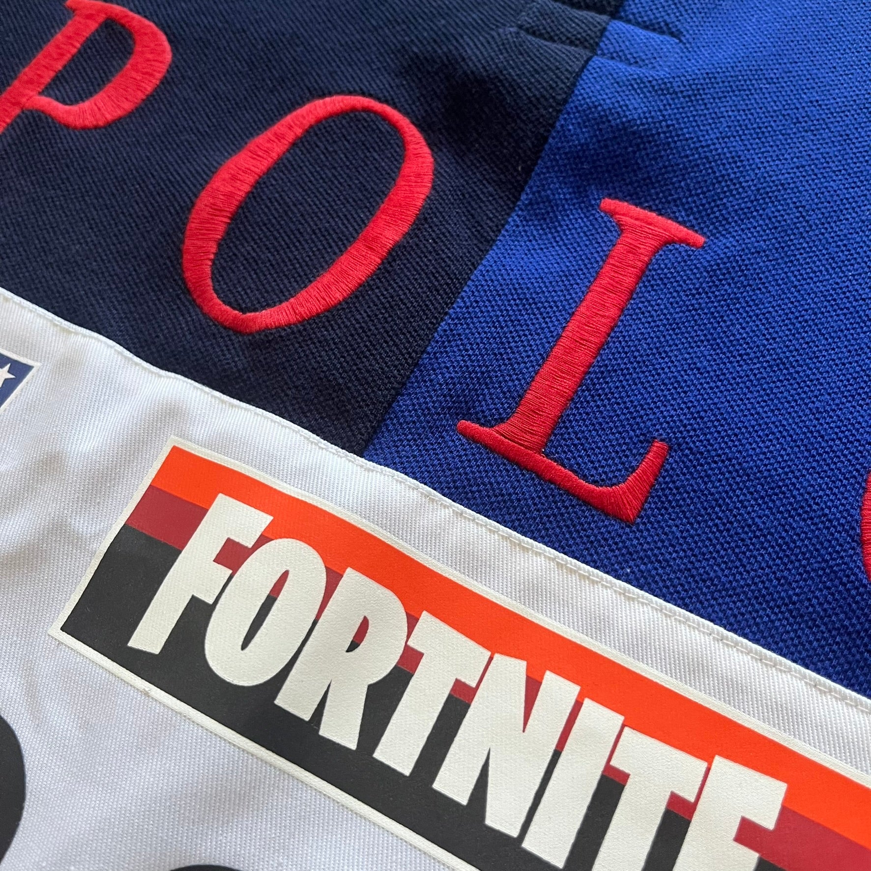 Polo Ralph Lauren x Fortnite 2022 Polo Shirt featuring a two-tone split design in blue and navy with a crisp white collar. The front showcases a large white panel with “2022” text, Ralph Lauren Est. 1967 branding, and a Fortnite logo, along with an oversized embroidered Polo logo above. A limited collaboration piece combining Ralph Lauren’s heritage aesthetic with Fortnite’s playful edge.