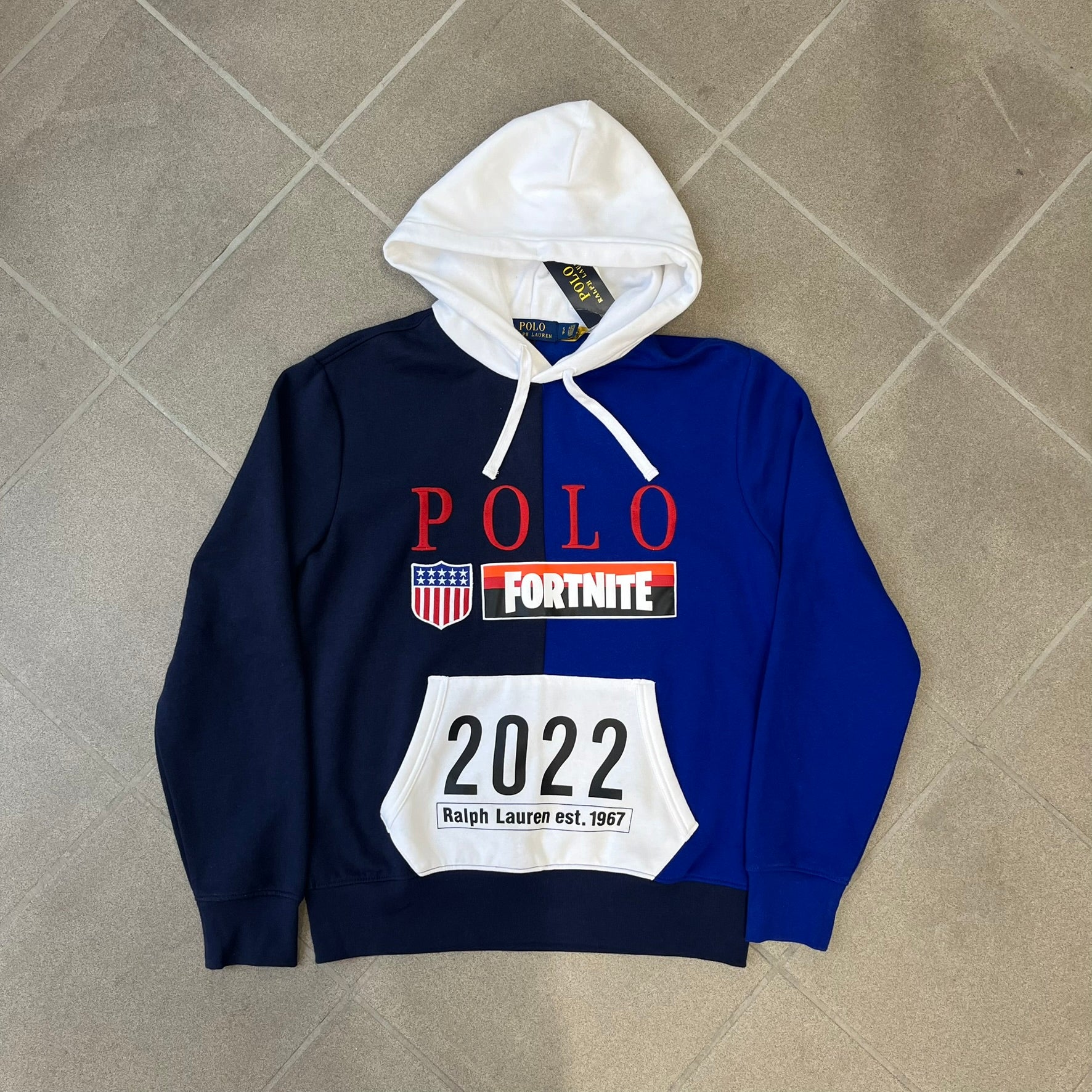 Polo Ralph Lauren x Fortnite 2022 Hoodie featuring a two-tone split design in blue and navy with a white hood. The front showcases a large white panel with “2022” text, Ralph Lauren Est. 1967 branding, and a Fortnite logo, along with an oversized embroidered Polo logo above. A limited collaboration piece with paper tags.