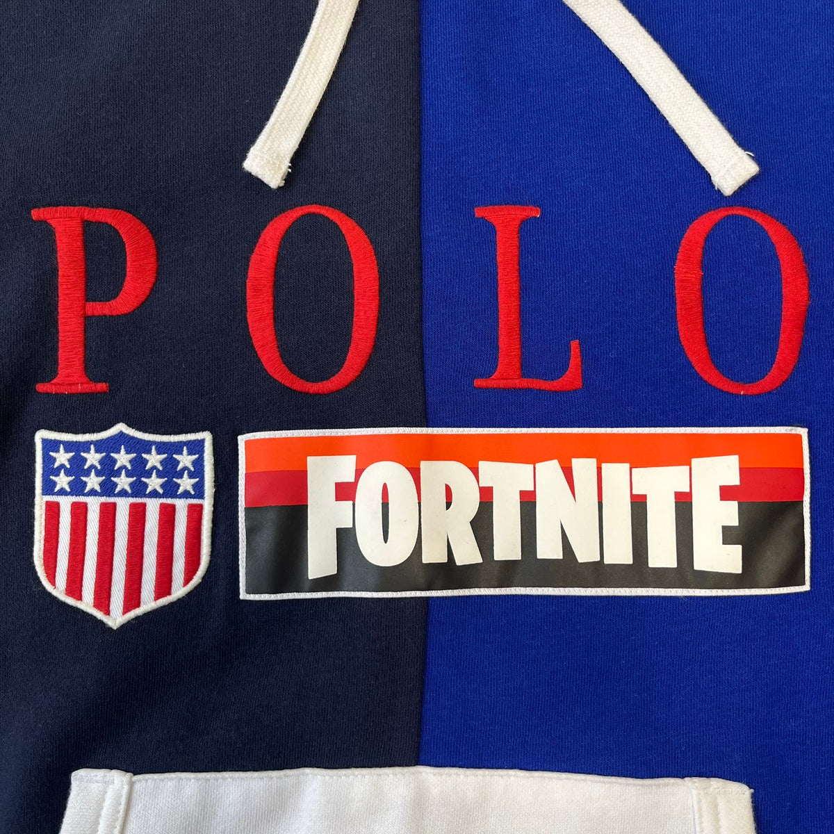 Polo Ralph Lauren x Fortnite 2022 Hoodie featuring a two-tone split design in blue and navy with a white hood. The front showcases a large white panel with “2022” text, Ralph Lauren Est. 1967 branding, and a Fortnite logo, along with an oversized embroidered Polo logo above. A limited collaboration piece with paper tags.
