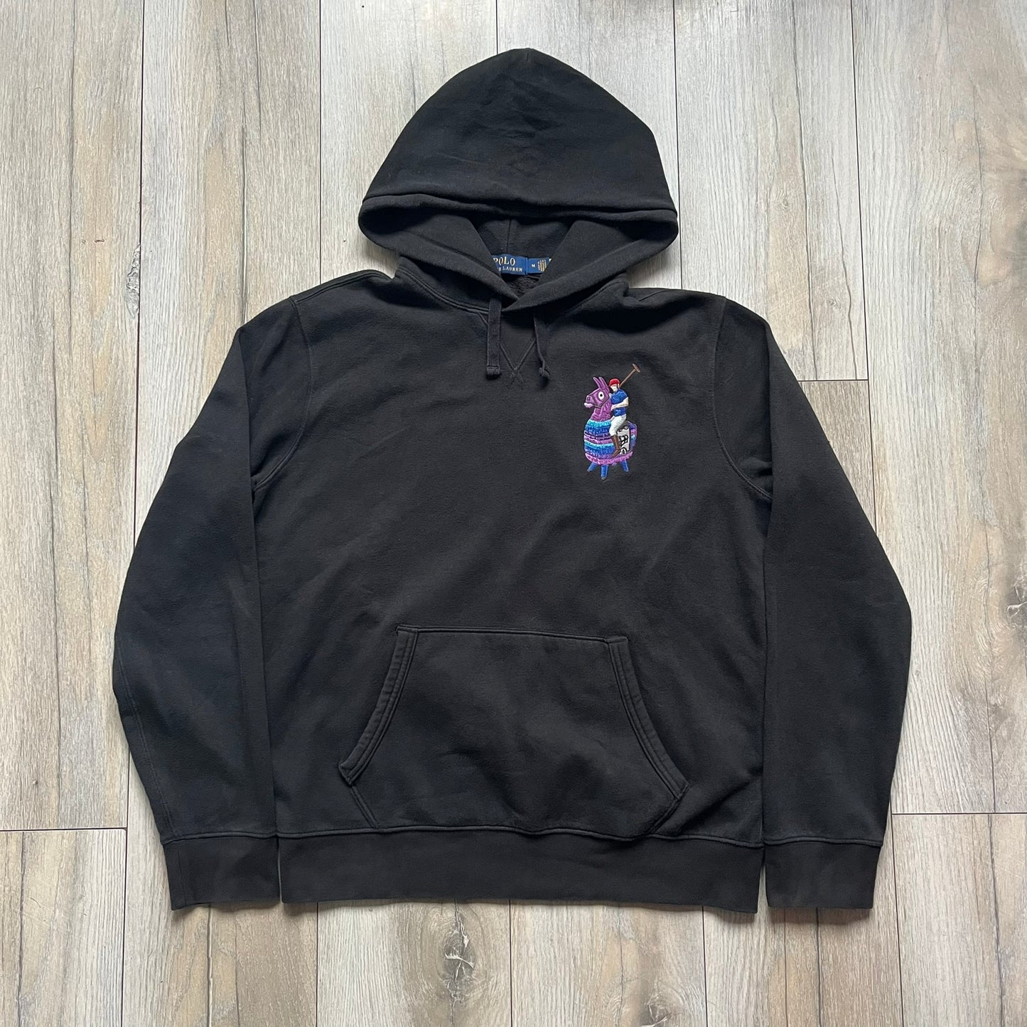 Polo Ralph Lauren x Fortnite black hoodie featuring a detailed embroidered purple Fortnite llama with the RL rider logo placed on top. Made from a 66% cotton and 34% polyester blend, it has a classic pullover construction with an attached hood and front kangaroo pocket. The embroidery is positioned on the left chest and stands out with clean stitching and sharp detail.