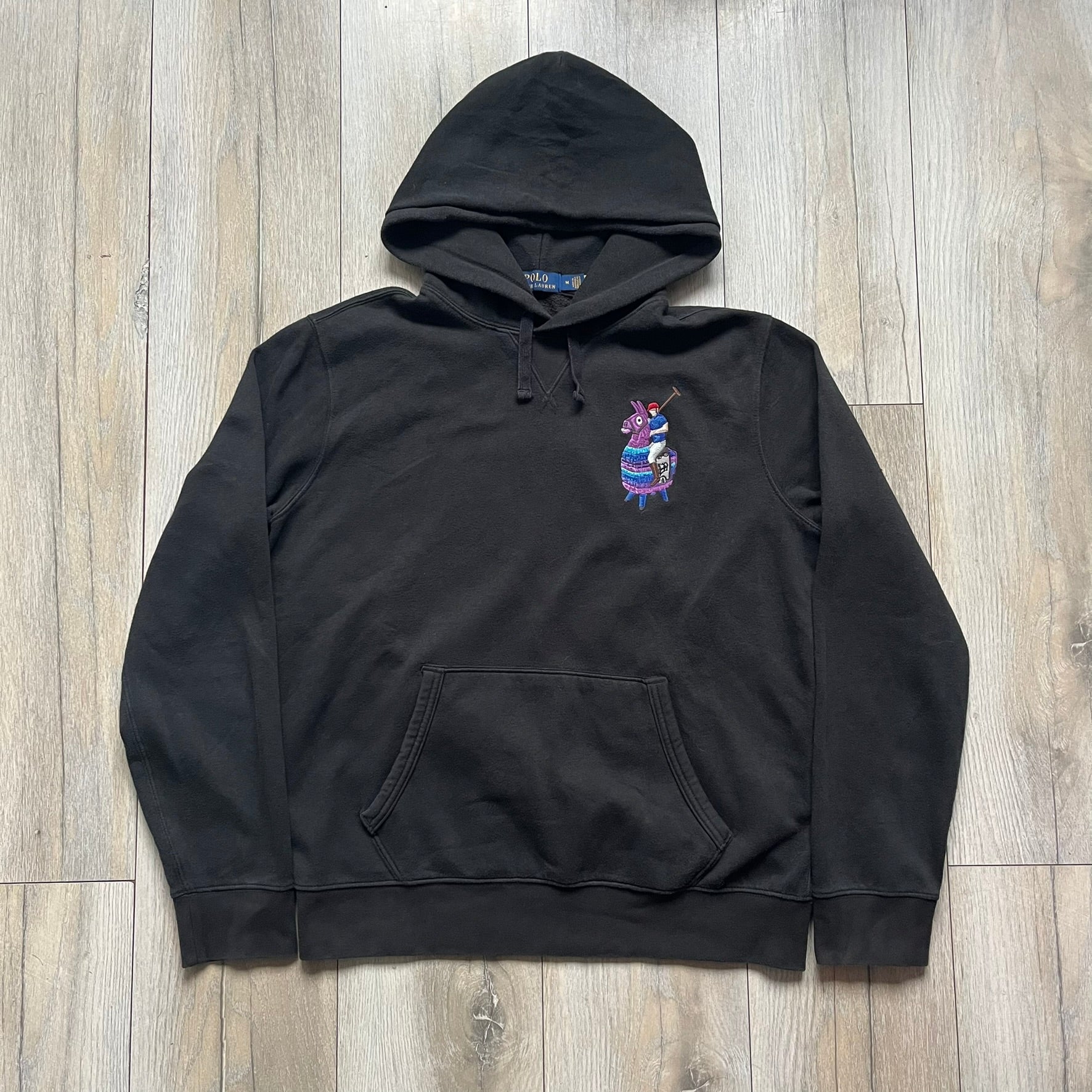 Polo Ralph Lauren x Fortnite black hoodie featuring a detailed embroidered purple Fortnite llama with the RL rider logo placed on top. Made from a 66% cotton and 34% polyester blend, it has a classic pullover construction with an attached hood and front kangaroo pocket. The embroidery is positioned on the left chest and stands out with clean stitching and sharp detail.