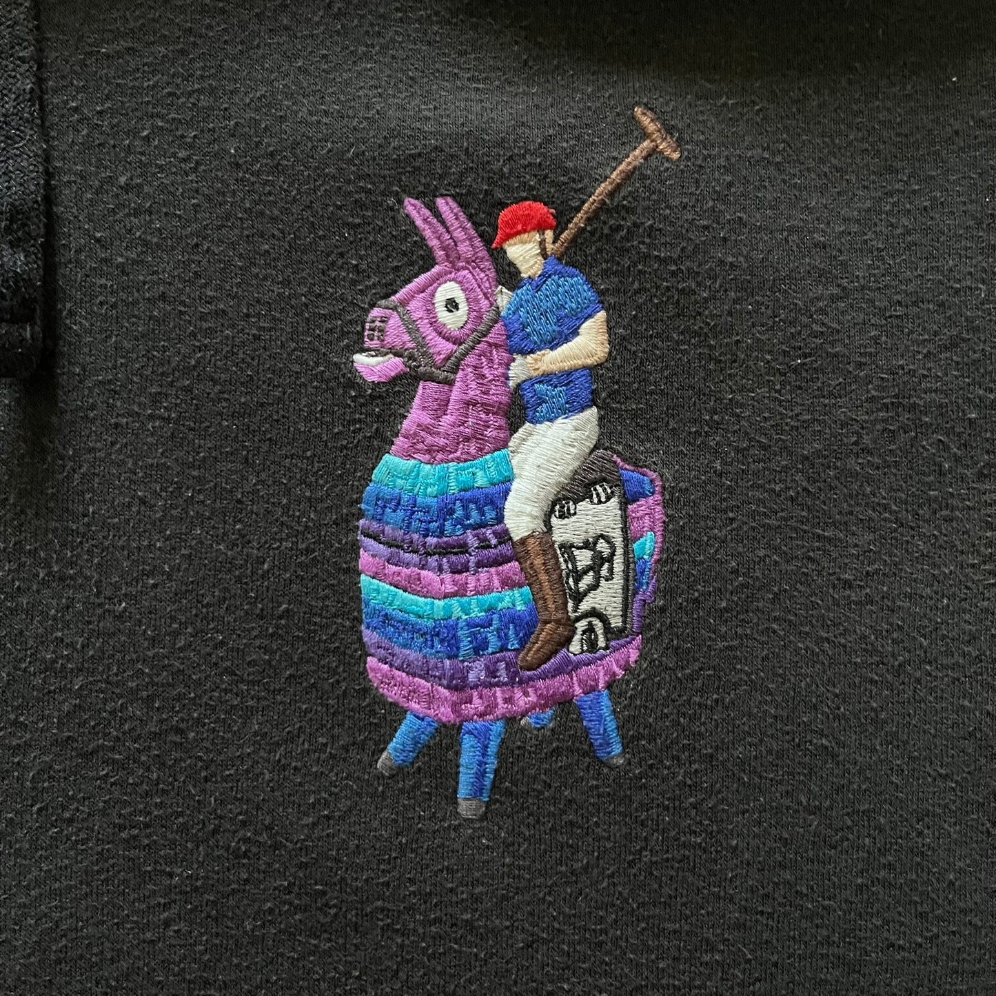  Polo Ralph Lauren x Fortnite black hoodie featuring a detailed embroidered purple Fortnite llama with the RL rider logo placed on top. Made from a 66% cotton and 34% polyester blend, it has a classic pullover construction with an attached hood and front kangaroo pocket. The embroidery is positioned on the left chest and stands out with clean stitching and sharp detail.