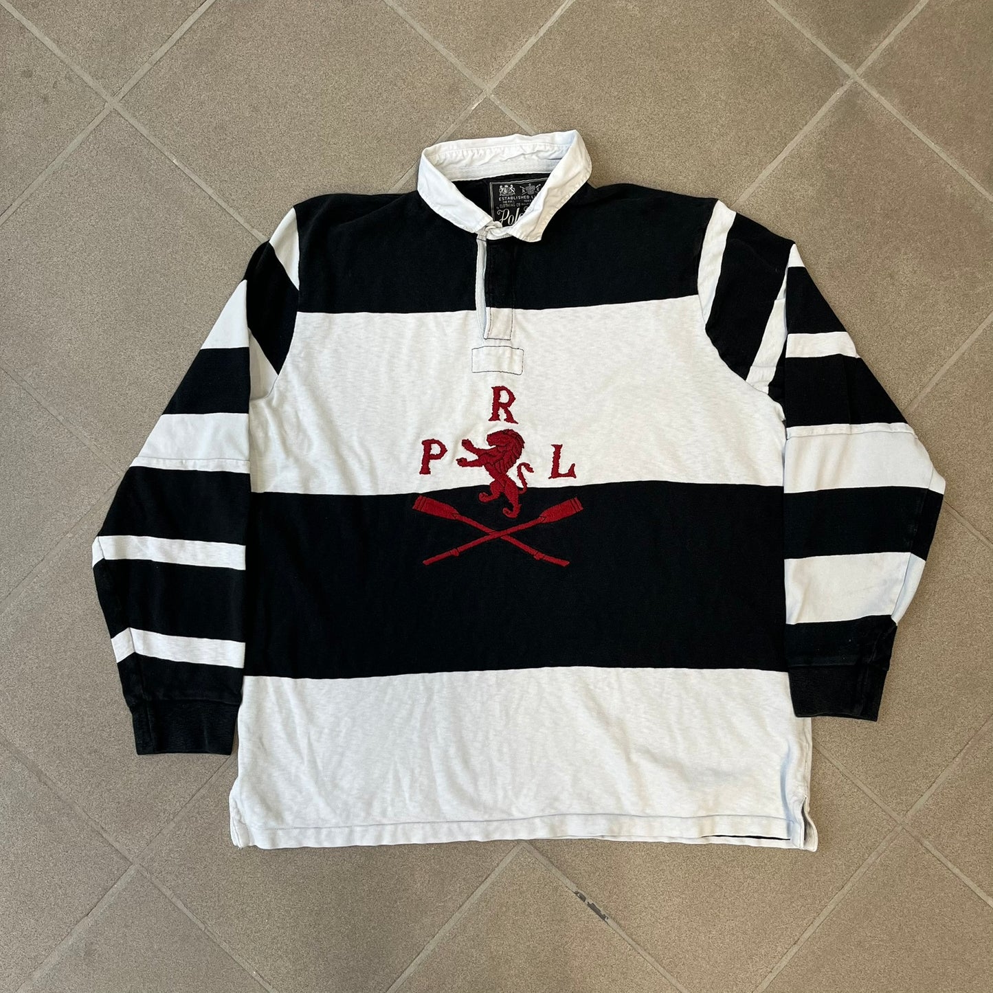 Polo Ralph Lauren Red Lion PRL Rugby Longsleeve features a black and white striped rugby design with four horizontal stripes across the front and additional stripes on the sleeves. The center of the chest has a red embroidered emblem featuring two crossed oars, a lion, and “PRL” lettering above the lion.