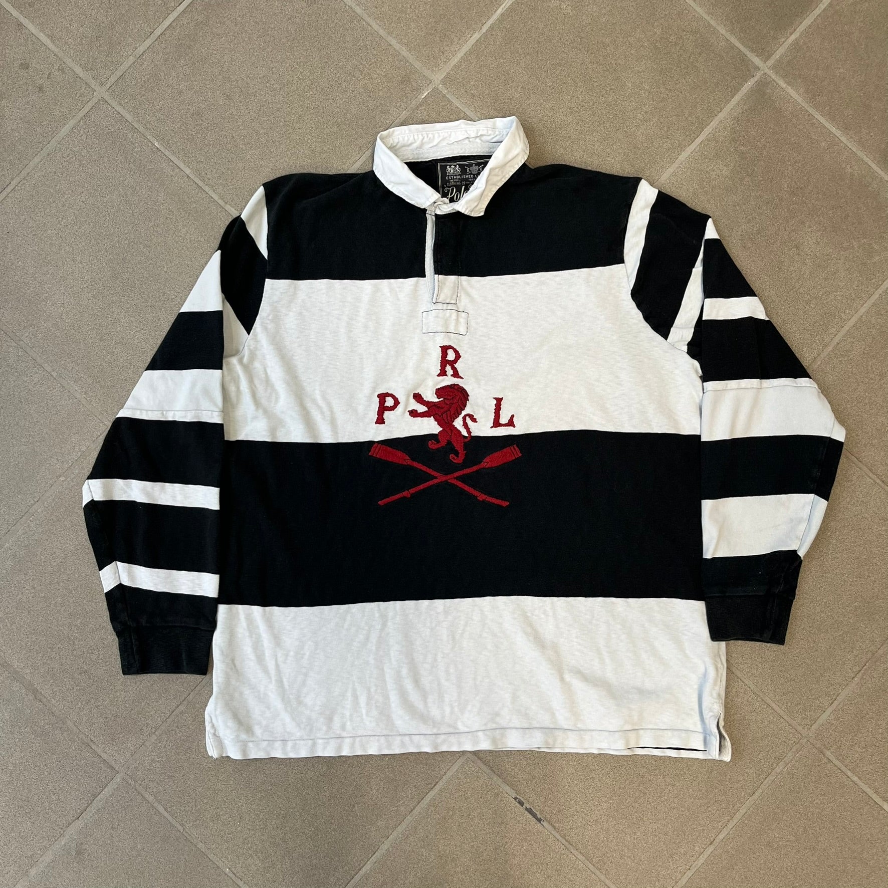 Polo Ralph Lauren Red Lion PRL Rugby Longsleeve features a black and white striped rugby design with four horizontal stripes across the front and additional stripes on the sleeves. The center of the chest has a red embroidered emblem featuring two crossed oars, a lion, and “PRL” lettering above the lion.