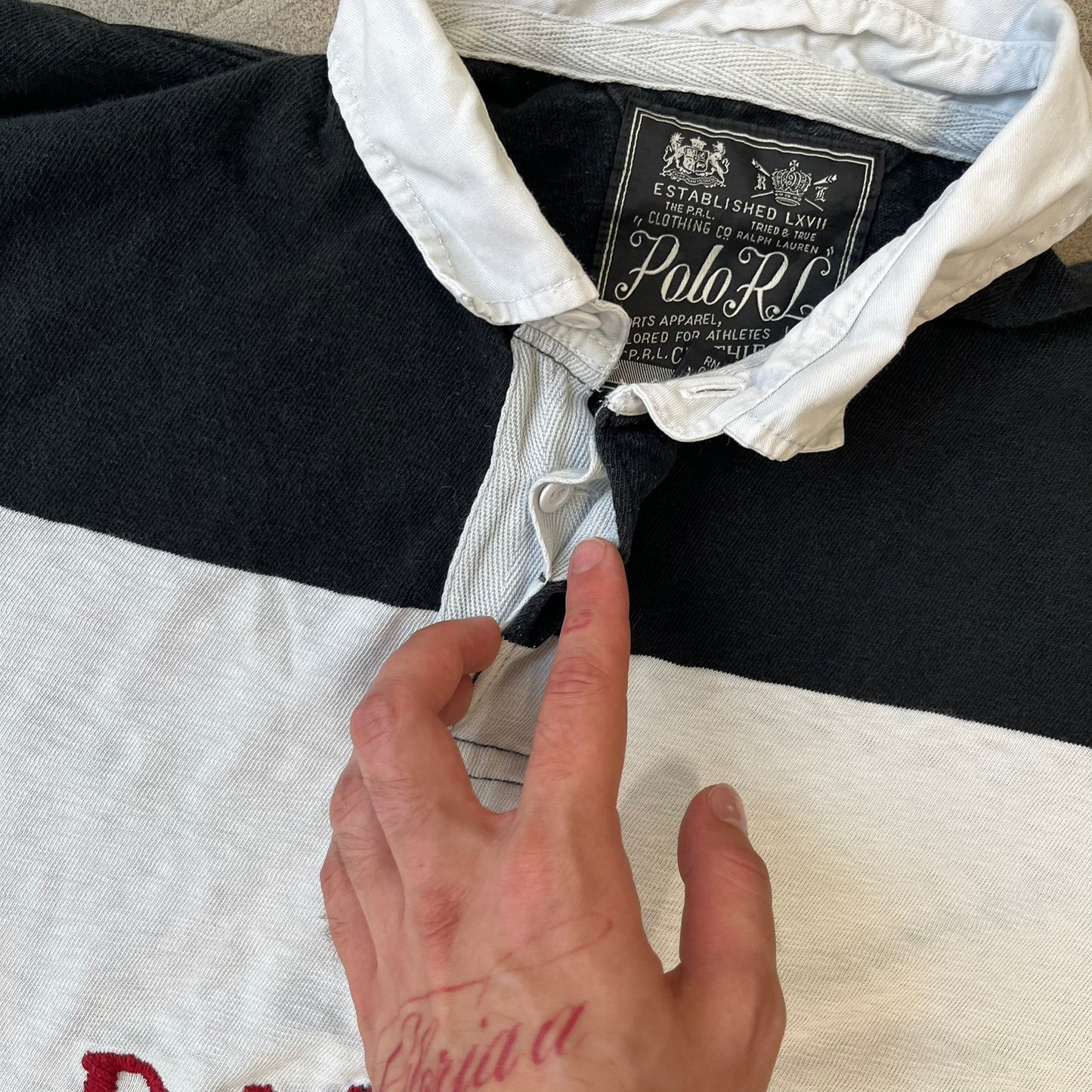 Polo Ralph Lauren Red Lion PRL Rugby Longsleeve features a black and white striped rugby design with four horizontal stripes across the front and additional stripes on the sleeves. The center of the chest has a red embroidered emblem featuring two crossed oars, a lion, and “PRL” lettering above the lion.