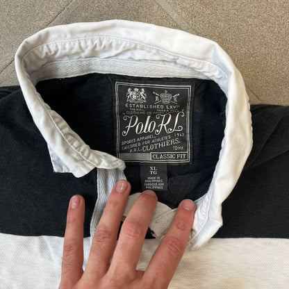 Polo Ralph Lauren Red Lion PRL Rugby Longsleeve features a black and white striped rugby design with four horizontal stripes across the front and additional stripes on the sleeves. The center of the chest has a red embroidered emblem featuring two crossed oars, a lion, and “PRL” lettering above the lion.
