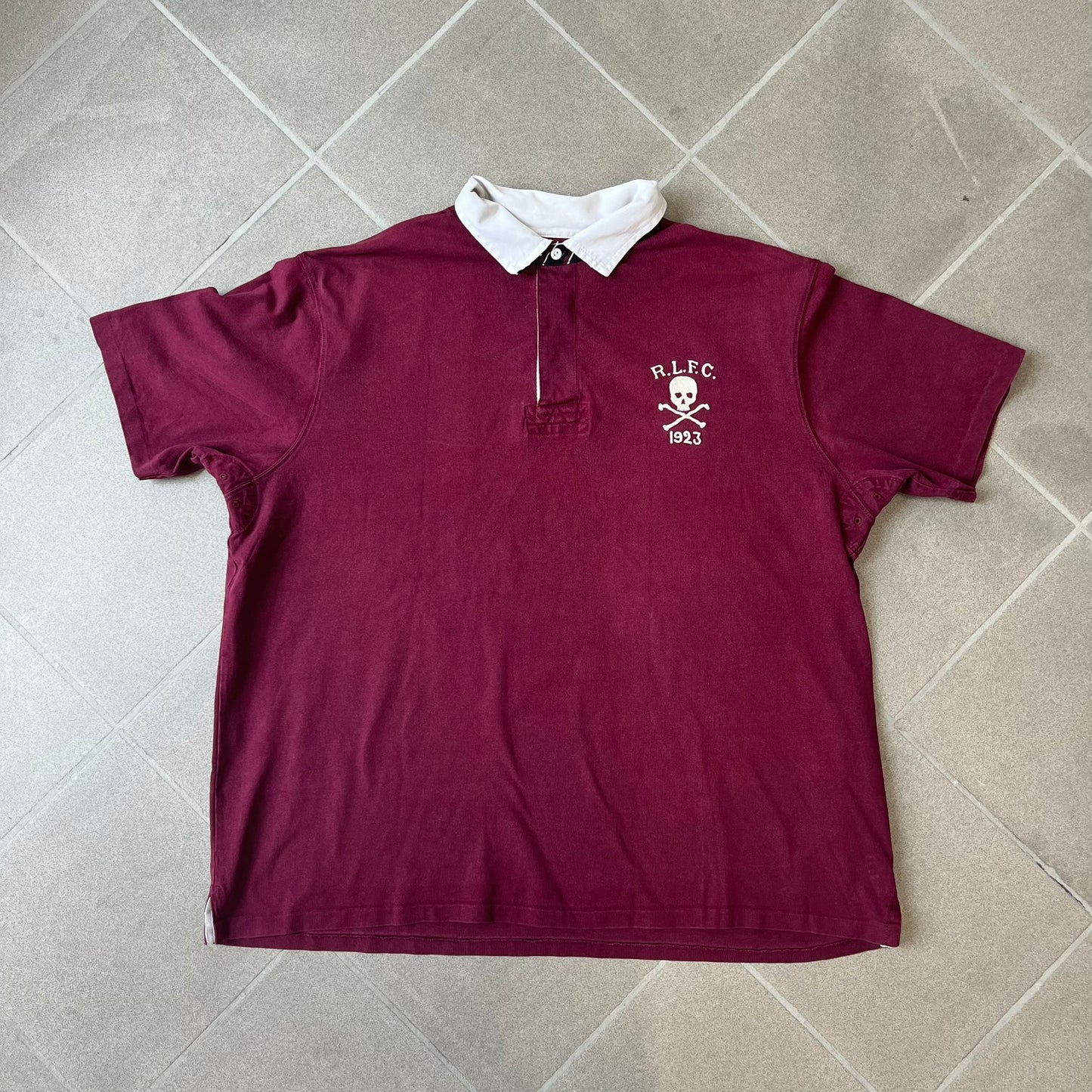 Polo Ralph Lauren “Skull &amp; Crossbone” Rugby Polo is a burgundy short-sleeve polo made from 100% cotton. It features a white contrasting rugby-style collar and a three-button placket. On the chest is a white embroidered skull and crossbones design with “1923” below it and “R.L.F.C” above. The design includes a split hem and embroidered detailing on the front.