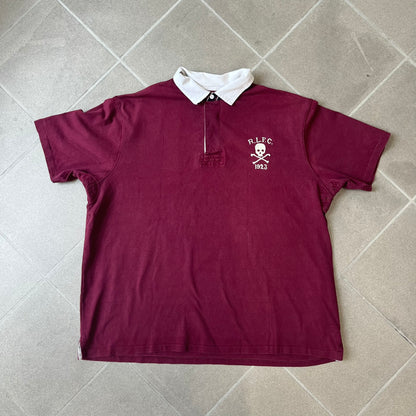 Polo Ralph Lauren “Skull &amp; Crossbone” Rugby Polo is a burgundy short-sleeve polo made from 100% cotton. It features a white contrasting rugby-style collar and a three-button placket. On the chest is a white embroidered skull and crossbones design with “1923” below it and “R.L.F.C” above. The design includes a split hem and embroidered detailing on the front.
