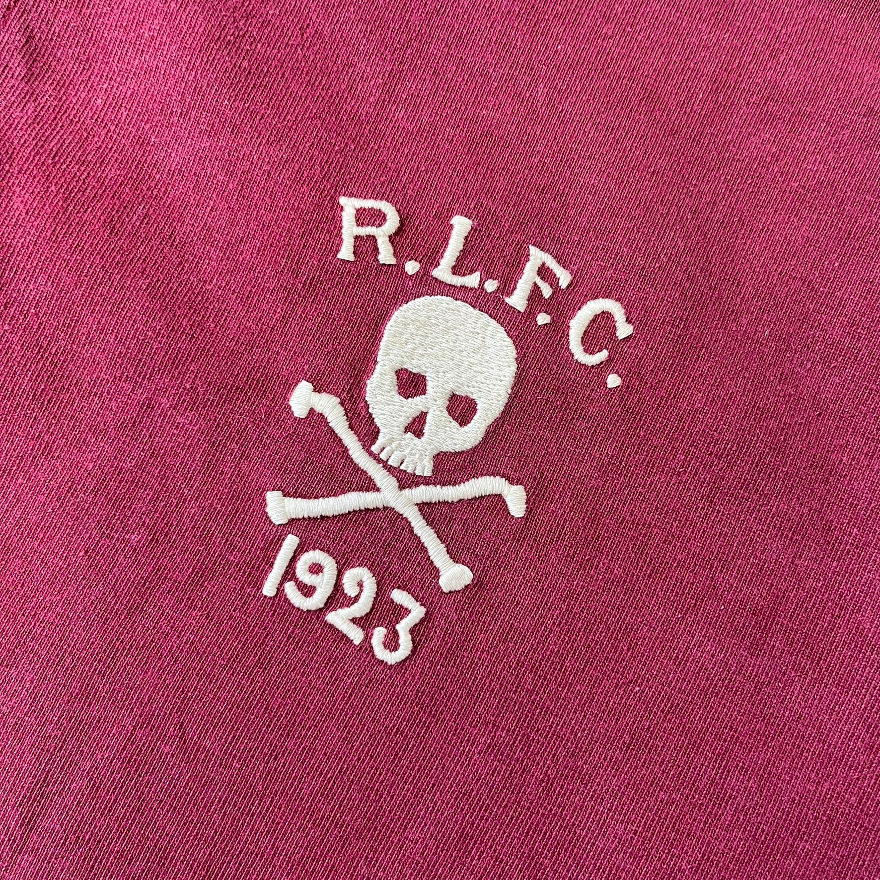Polo Ralph Lauren “Skull &amp; Crossbone” Rugby Polo is a burgundy short-sleeve polo made from 100% cotton. It features a white contrasting rugby-style collar and a three-button placket. On the chest is a white embroidered skull and crossbones design with “1923” below it and “R.L.F.C” above. The design includes a split hem and embroidered detailing on the front.