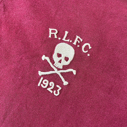 Polo Ralph Lauren “Skull &amp; Crossbone” Rugby Polo is a burgundy short-sleeve polo made from 100% cotton. It features a white contrasting rugby-style collar and a three-button placket. On the chest is a white embroidered skull and crossbones design with “1923” below it and “R.L.F.C” above. The design includes a split hem and embroidered detailing on the front.