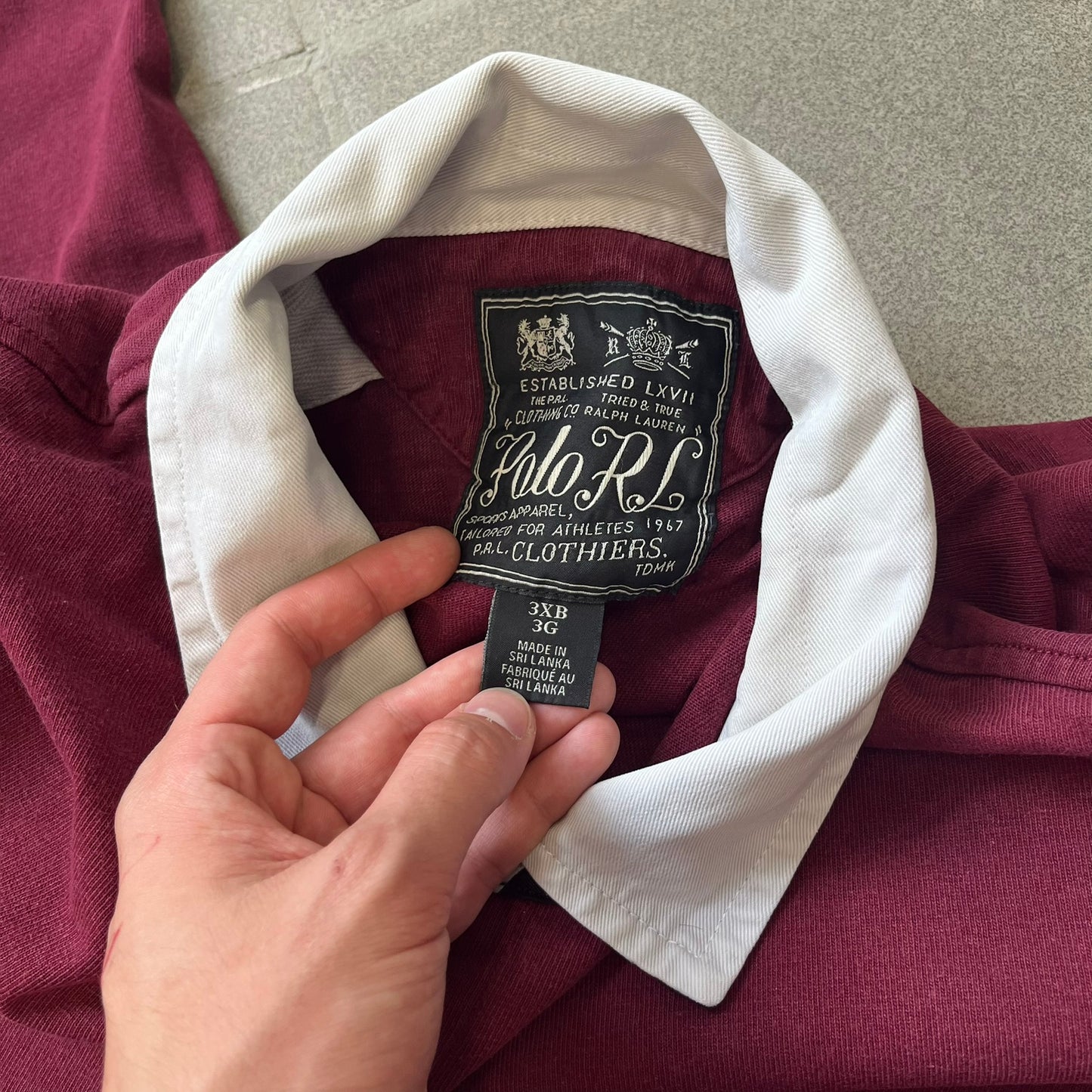 Polo Ralph Lauren “Skull &amp; Crossbone” Rugby Polo is a burgundy short-sleeve polo made from 100% cotton. It features a white contrasting rugby-style collar and a three-button placket. On the chest is a white embroidered skull and crossbones design with “1923” below it and “R.L.F.C” above. The design includes a split hem and embroidered detailing on the front.