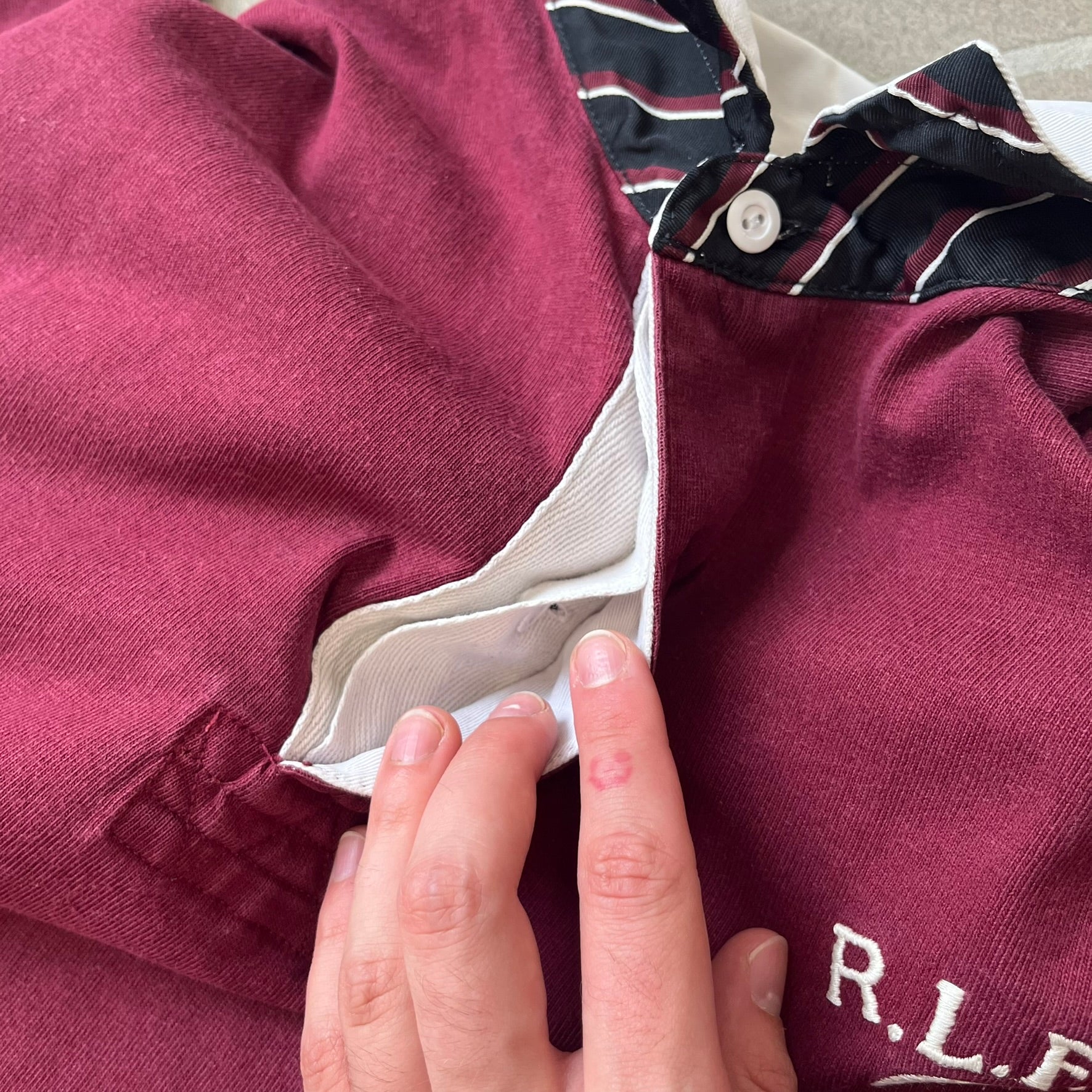 Polo Ralph Lauren “Skull &amp; Crossbone” Rugby Polo is a burgundy short-sleeve polo made from 100% cotton. It features a white contrasting rugby-style collar and a three-button placket. On the chest is a white embroidered skull and crossbones design with “1923” below it and “R.L.F.C” above. The design includes a split hem and embroidered detailing on the front.