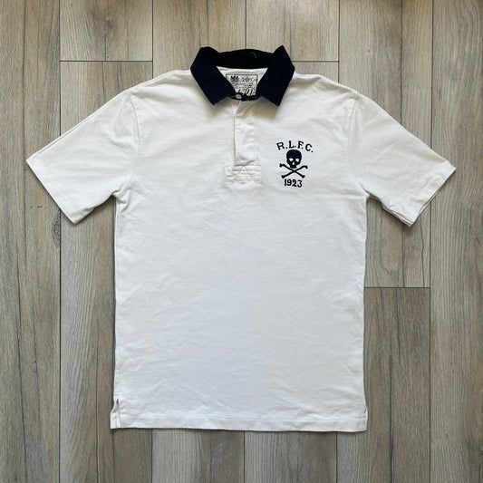 Polo Ralph Lauren “Skull &amp; Crossbone” Rugby Polo is a white short-sleeve polo made from 100% cotton. It features a black contrasting rugby-style collar and a three-button placket. On the chest is a black embroidered skull and crossbones design with “1923” below it and “R.L.F.C” above. The design includes a split hem and embroidered detailing on the front.