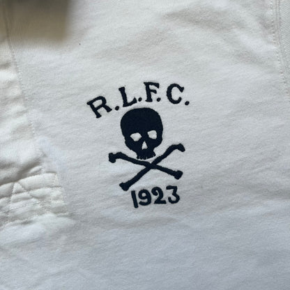 Polo Ralph Lauren “Skull &amp; Crossbone” Rugby Polo is a white short-sleeve polo made from 100% cotton. It features a black contrasting rugby-style collar and a three-button placket. On the chest is a black embroidered skull and crossbones design with “1923” below it and “R.L.F.C” above. The design includes a split hem and embroidered detailing on the front.