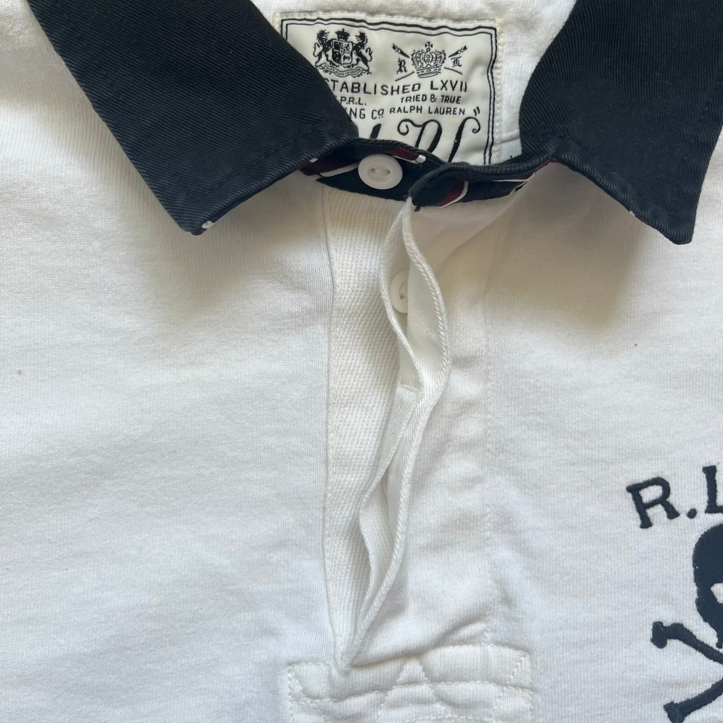Polo Ralph Lauren “Skull &amp; Crossbone” Rugby Polo is a white short-sleeve polo made from 100% cotton. It features a black contrasting rugby-style collar and a three-button placket. On the chest is a black embroidered skull and crossbones design with “1923” below it and “R.L.F.C” above. The design includes a split hem and embroidered detailing on the front.