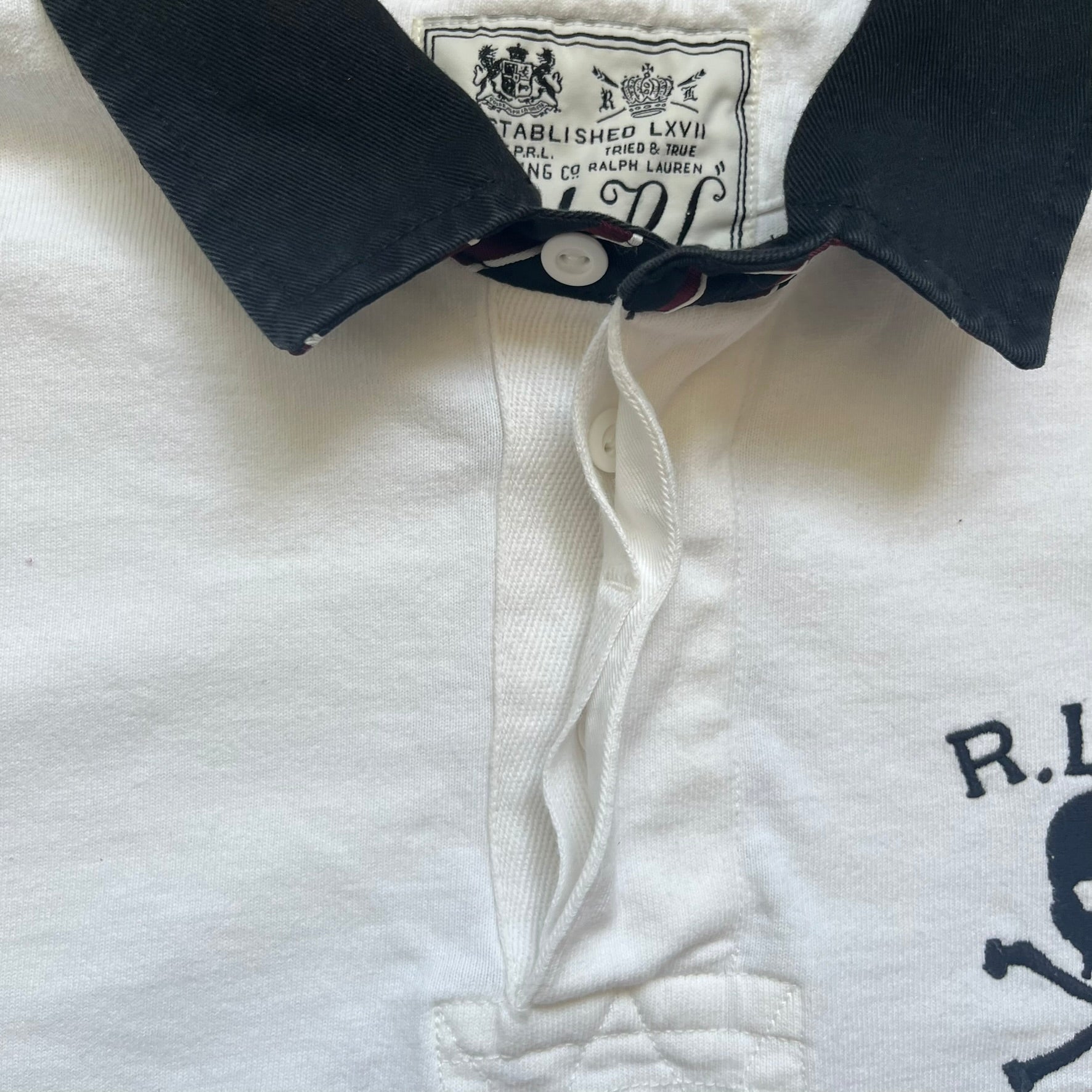 Polo Ralph Lauren “Skull &amp; Crossbone” Rugby Polo is a white short-sleeve polo made from 100% cotton. It features a black contrasting rugby-style collar and a three-button placket. On the chest is a black embroidered skull and crossbones design with “1923” below it and “R.L.F.C” above. The design includes a split hem and embroidered detailing on the front.