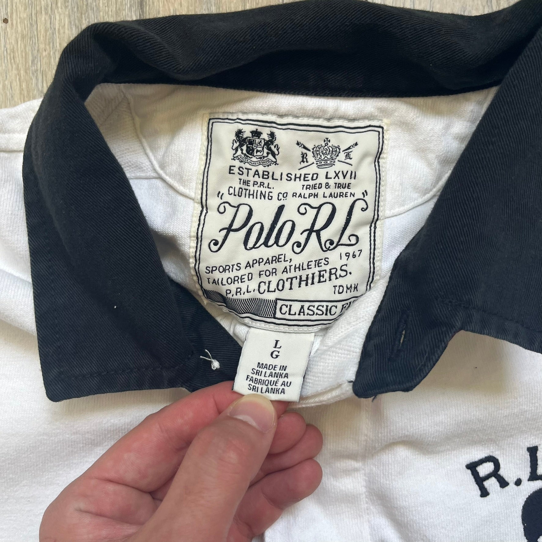 Polo Ralph Lauren “Skull &amp; Crossbone” Rugby Polo is a white short-sleeve polo made from 100% cotton. It features a black contrasting rugby-style collar and a three-button placket. On the chest is a black embroidered skull and crossbones design with “1923” below it and “R.L.F.C” above. The design includes a split hem and embroidered detailing on the front.