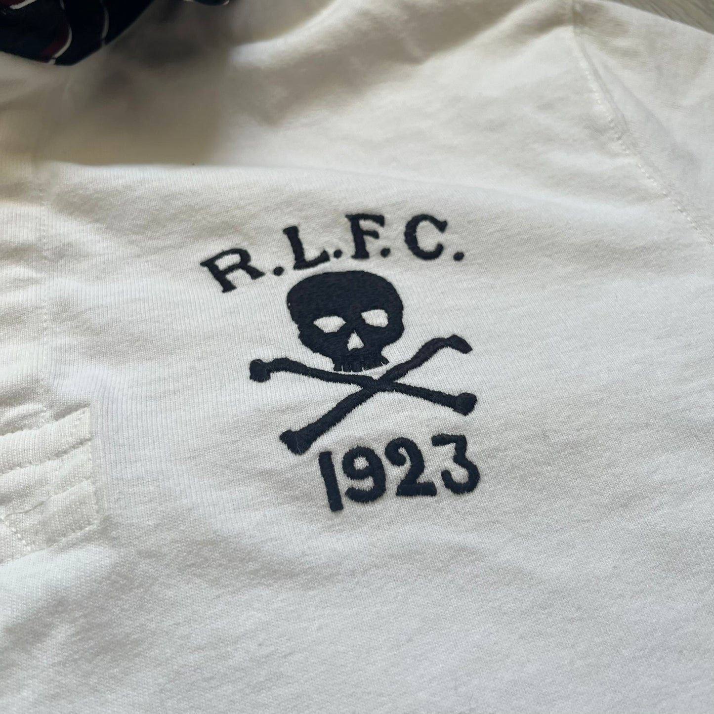 Polo Ralph Lauren “Skull &amp; Crossbone” Rugby Polo is a white short-sleeve polo made from 100% cotton. It features a black contrasting rugby-style collar and a three-button placket. On the chest is a black embroidered skull and crossbones design with “1923” below it and “R.L.F.C” above. The design includes a split hem and embroidered detailing on the front.