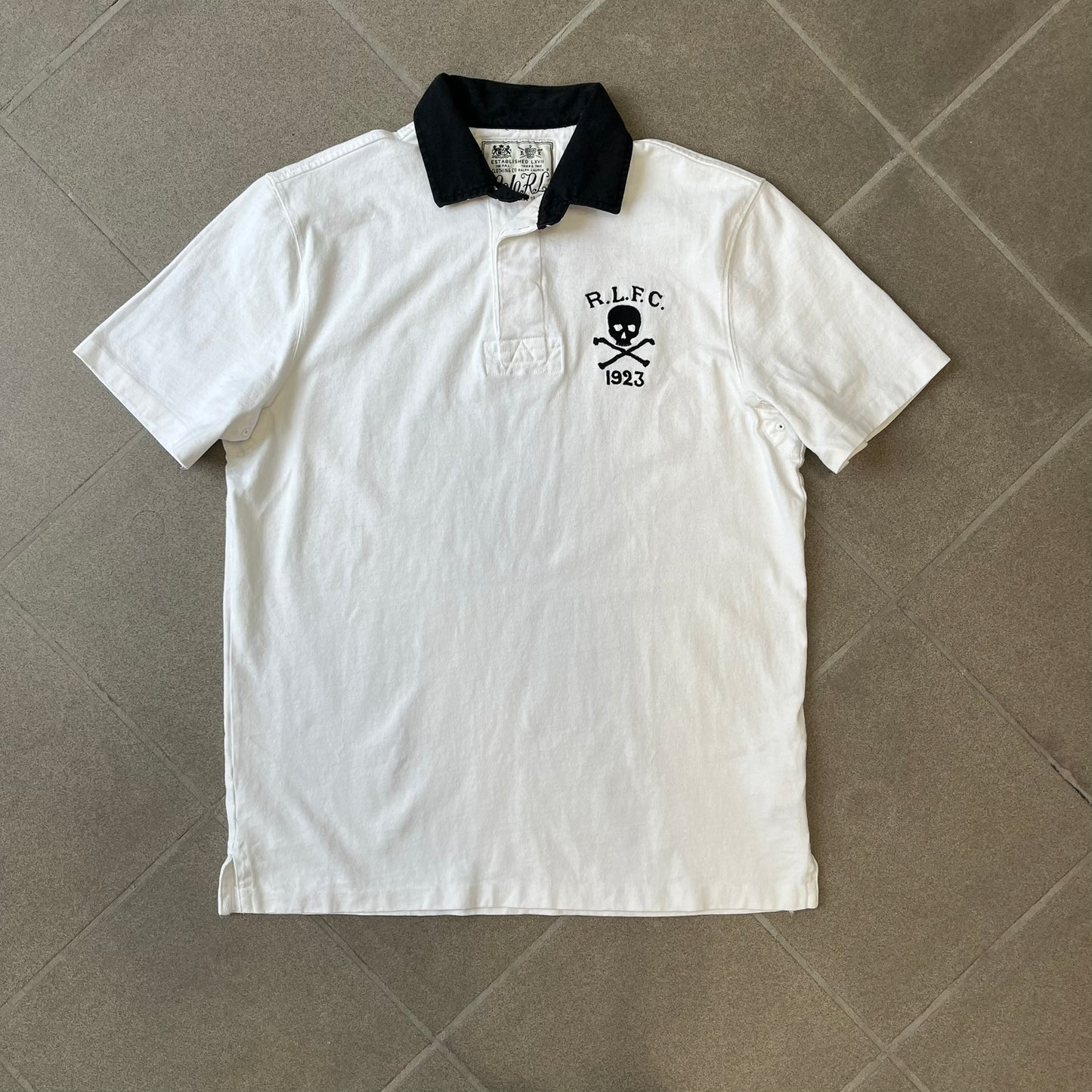 Polo Ralph Lauren “Skull &amp; Crossbone” Rugby Polo is a white short-sleeve polo made from 100% cotton. It features a black contrasting rugby-style collar and a three-button placket. On the chest is a black embroidered skull and crossbones design with “1923” below it and “R.L.F.C” above. The design includes a split hem and embroidered detailing on the front.