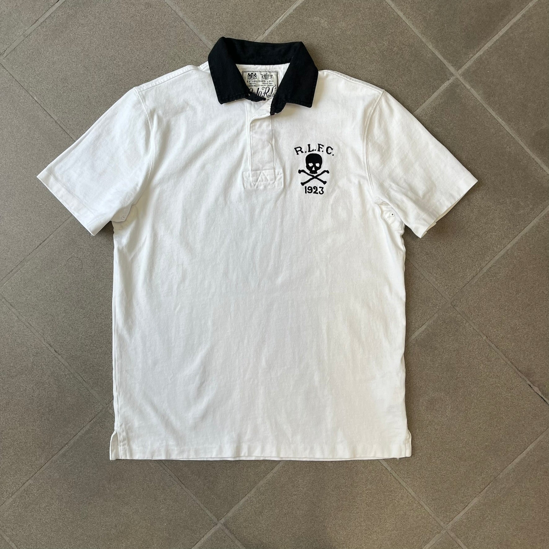 Polo Ralph Lauren “Skull &amp; Crossbone” Rugby Polo is a white short-sleeve polo made from 100% cotton. It features a black contrasting rugby-style collar and a three-button placket. On the chest is a black embroidered skull and crossbones design with “1923” below it and “R.L.F.C” above. The design includes a split hem and embroidered detailing on the front.