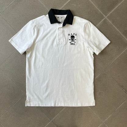 Polo Ralph Lauren “Skull &amp; Crossbone” Rugby Polo is a white short-sleeve polo made from 100% cotton. It features a black contrasting rugby-style collar and a three-button placket. On the chest is a black embroidered skull and crossbones design with “1923” below it and “R.L.F.C” above. The design includes a split hem and embroidered detailing on the front.