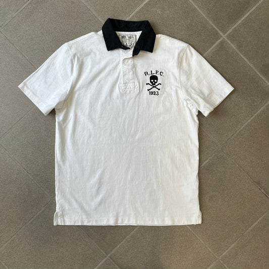 Polo Ralph Lauren “Skull &amp; Crossbone” Rugby Polo is a white short-sleeve polo made from 100% cotton. It features a black contrasting rugby-style collar and a three-button placket. On the chest is a black embroidered skull and crossbones design with “1923” below it and “R.L.F.C” above. The design includes a split hem and embroidered detailing on the front.